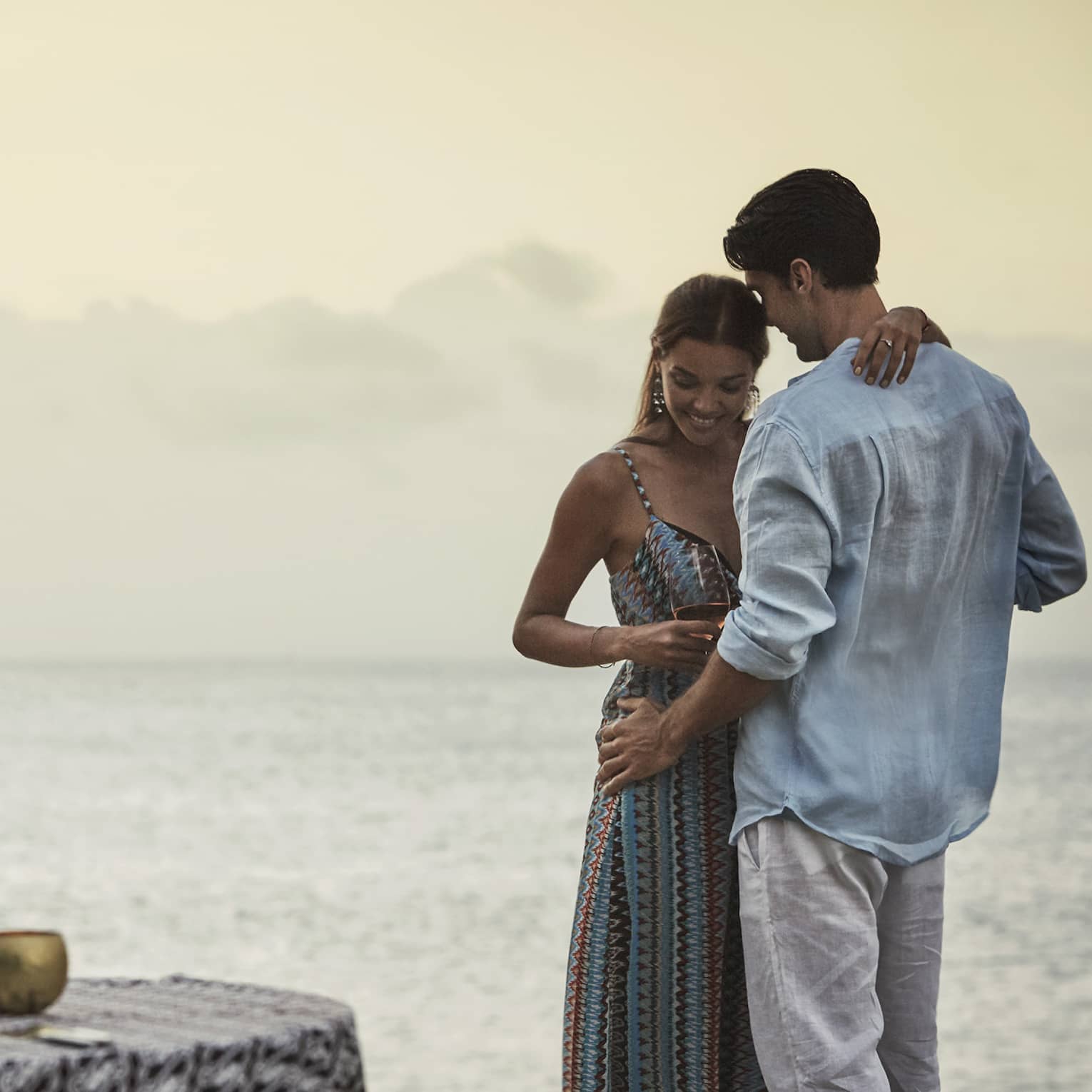Couple embraces beside private dining table overlooking ocean at sunset