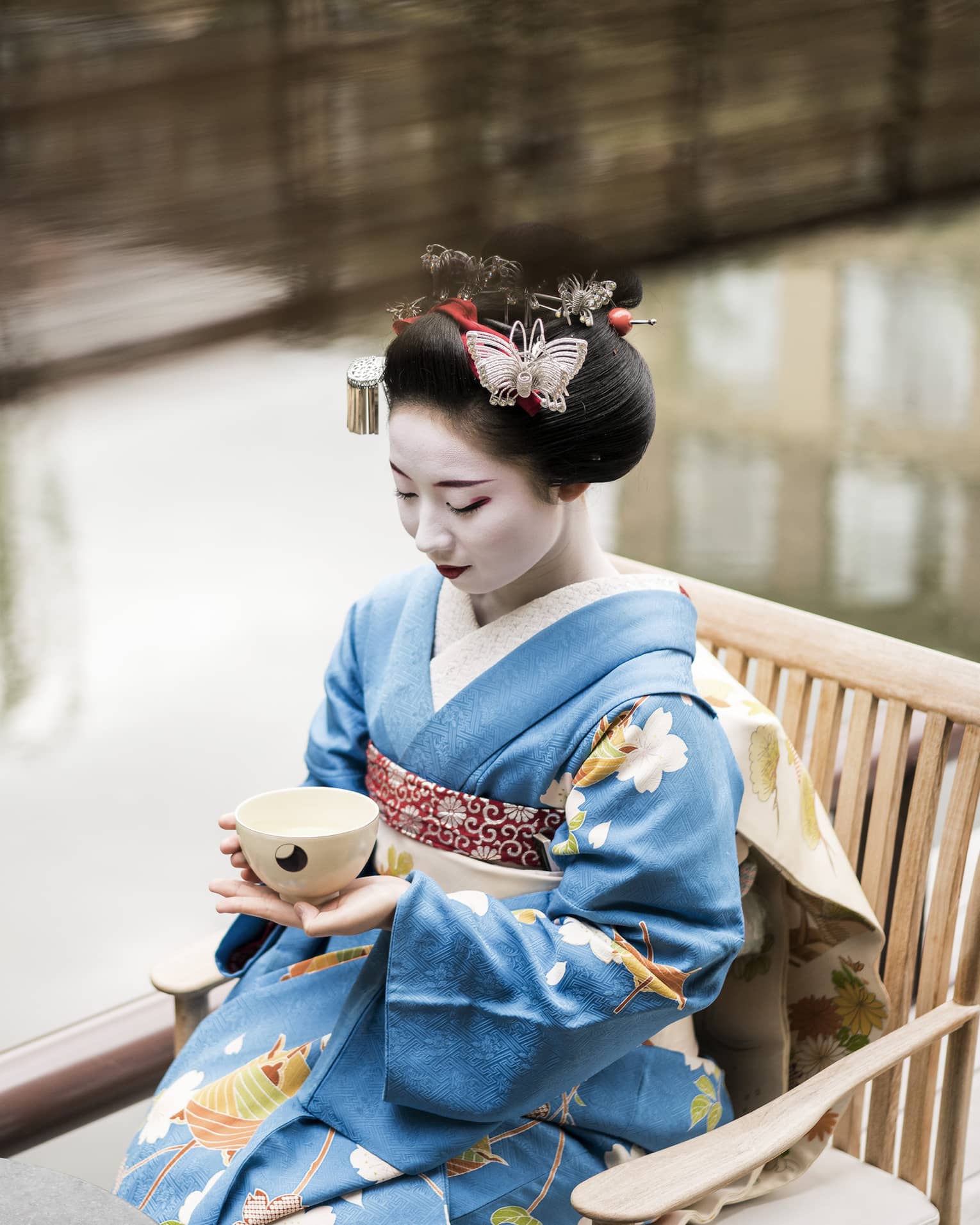 Geisha wearing traditional kimono robe, makeup sits on wood chair, holds tea cup