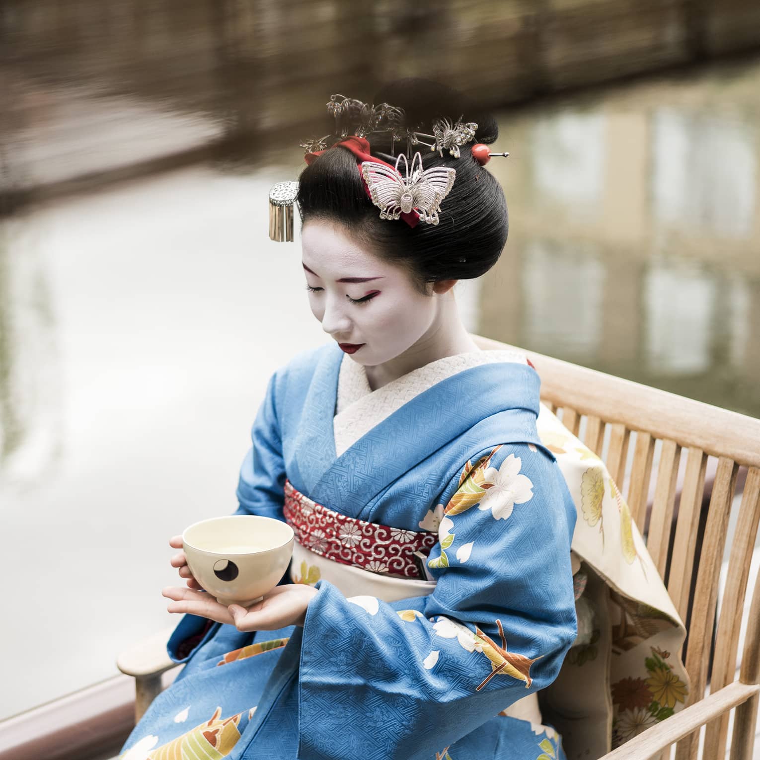 Geisha wearing traditional kimono robe, makeup sits on wood chair, holds tea cup
