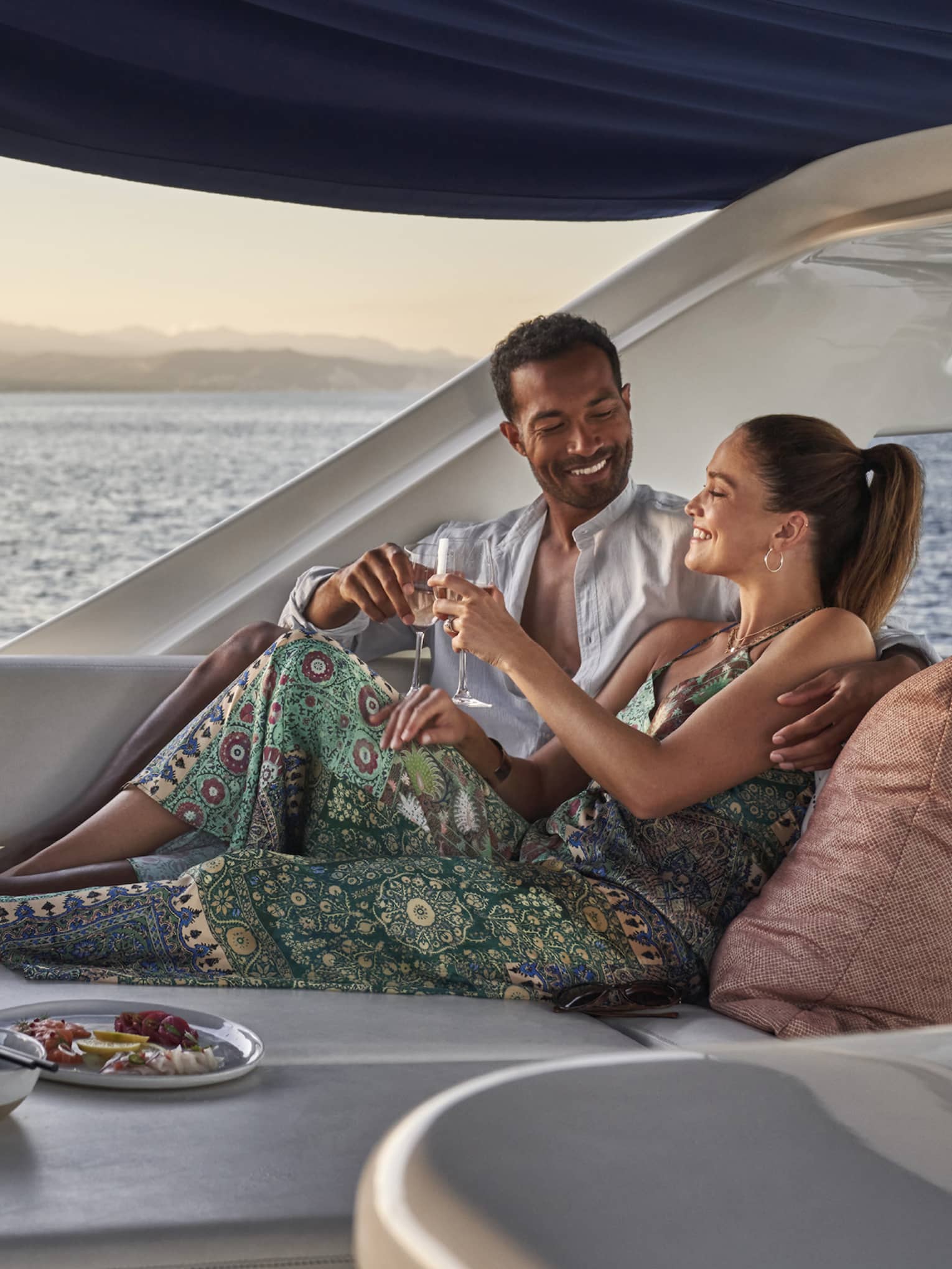 Aboard a boat in open air under a sunshade, a cuddling couple clink wine glasses amid cushions, dishes and an ice bucket.