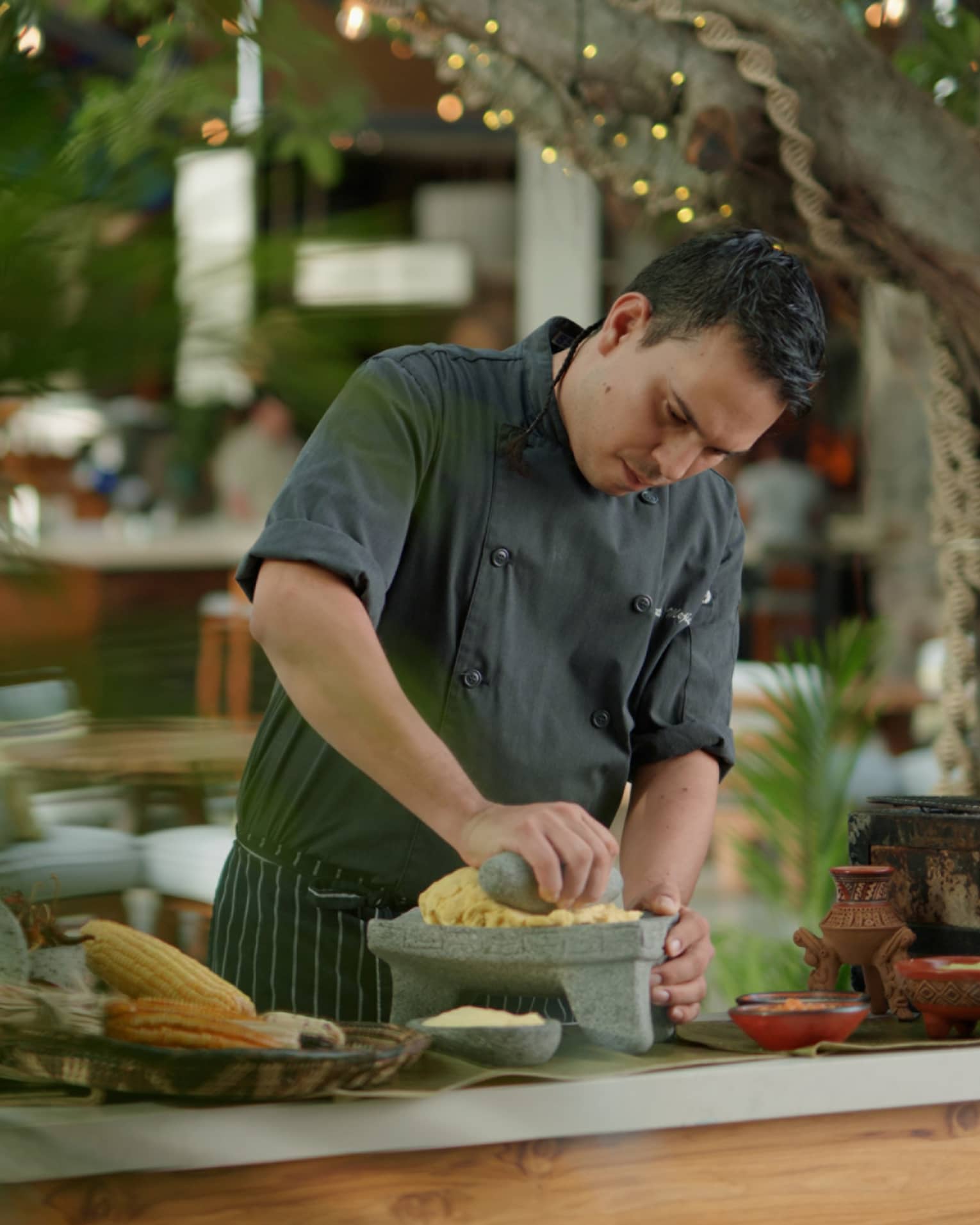 Chef wearing dark grey uniform mashes corn using a stone mortar and pestle