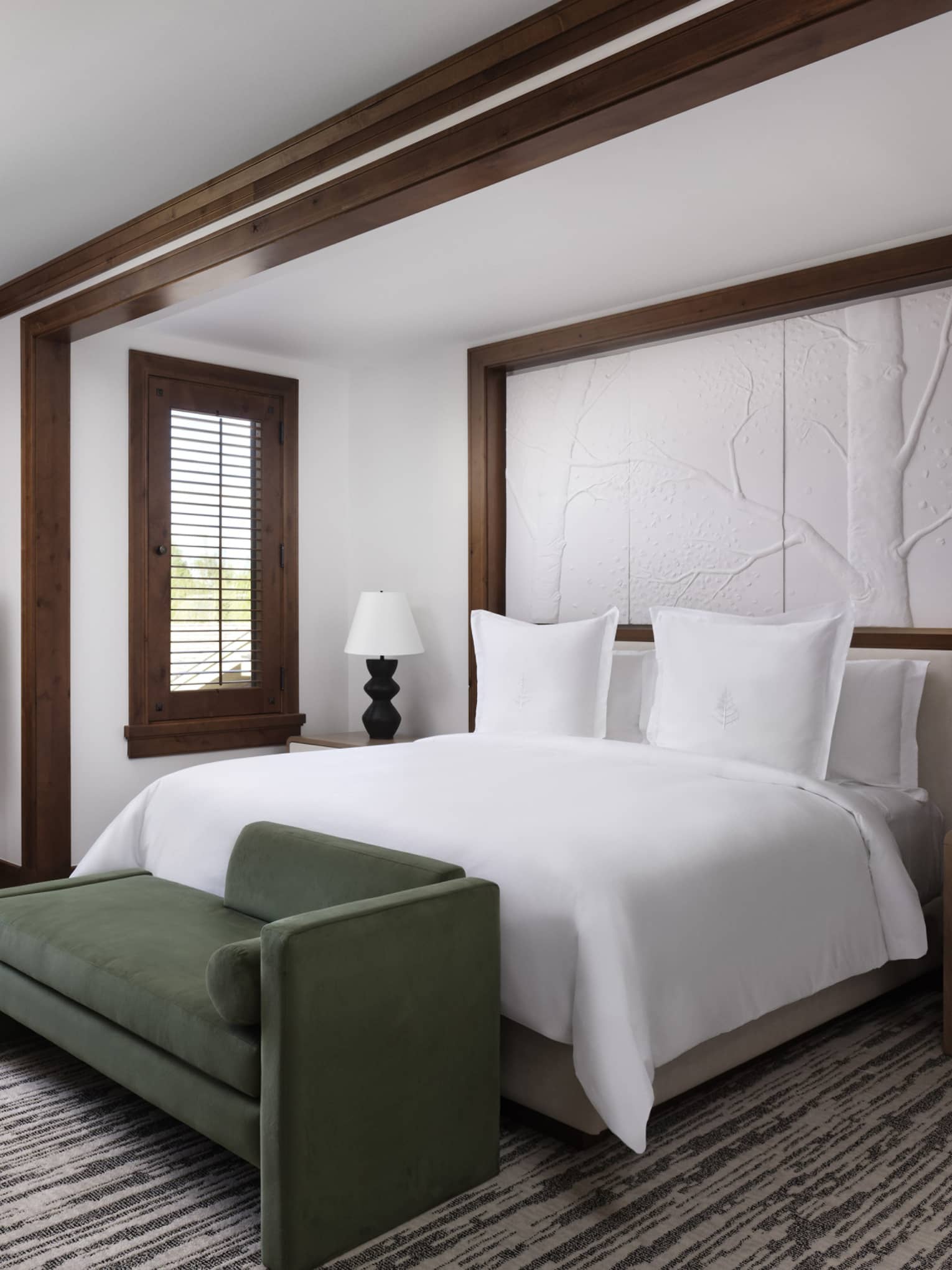 Luxury resort room with a king bed, wooden accents, dark green bench, nightstands with lamps, table with two chairs, and large windows overlooking the valley