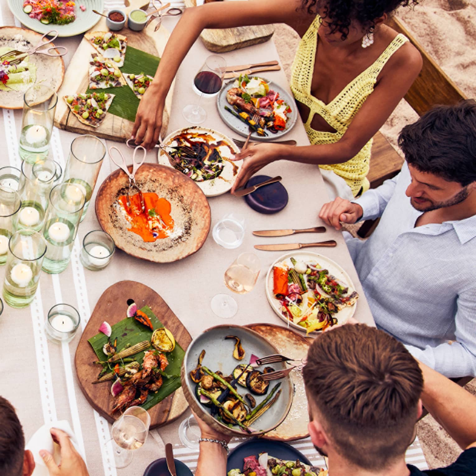 Beachside dining table with an assortment of vibrant dishes, candlelit glass holders and guests enjoying fresh seafood and grilled vegetables