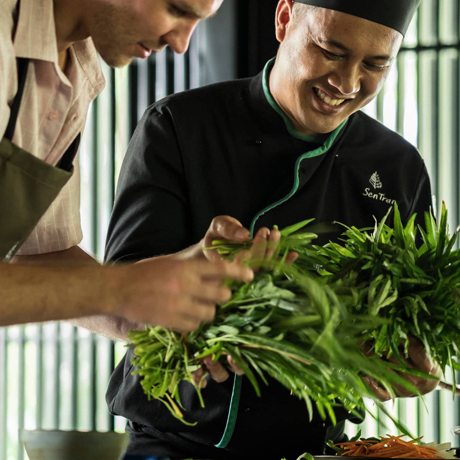 Smiling Chef Tran Van Sen holds greens, shows them to man in apron