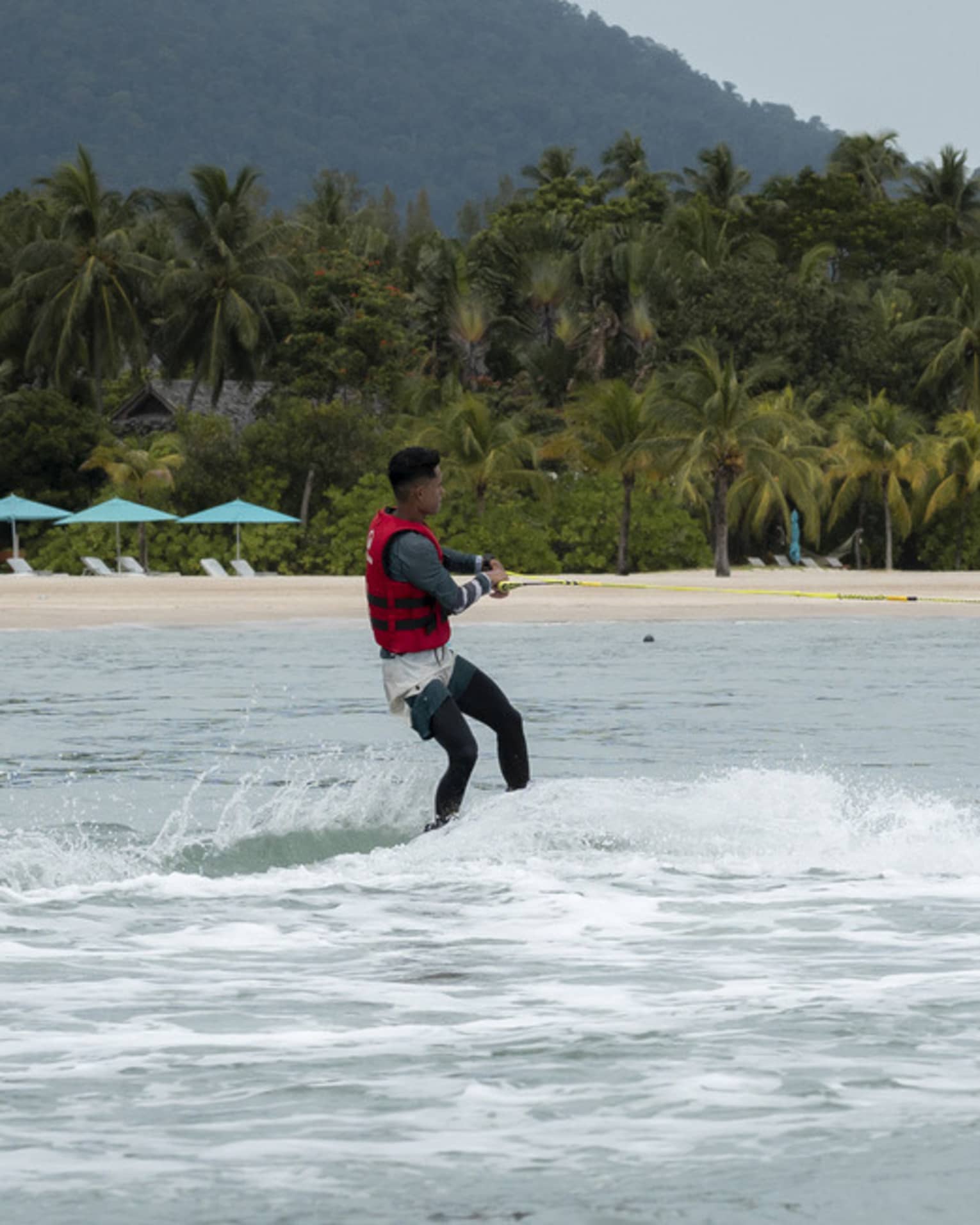 Person in red life jacket glides along the water on a water ski, the shore and copse of palm trees in the distance
