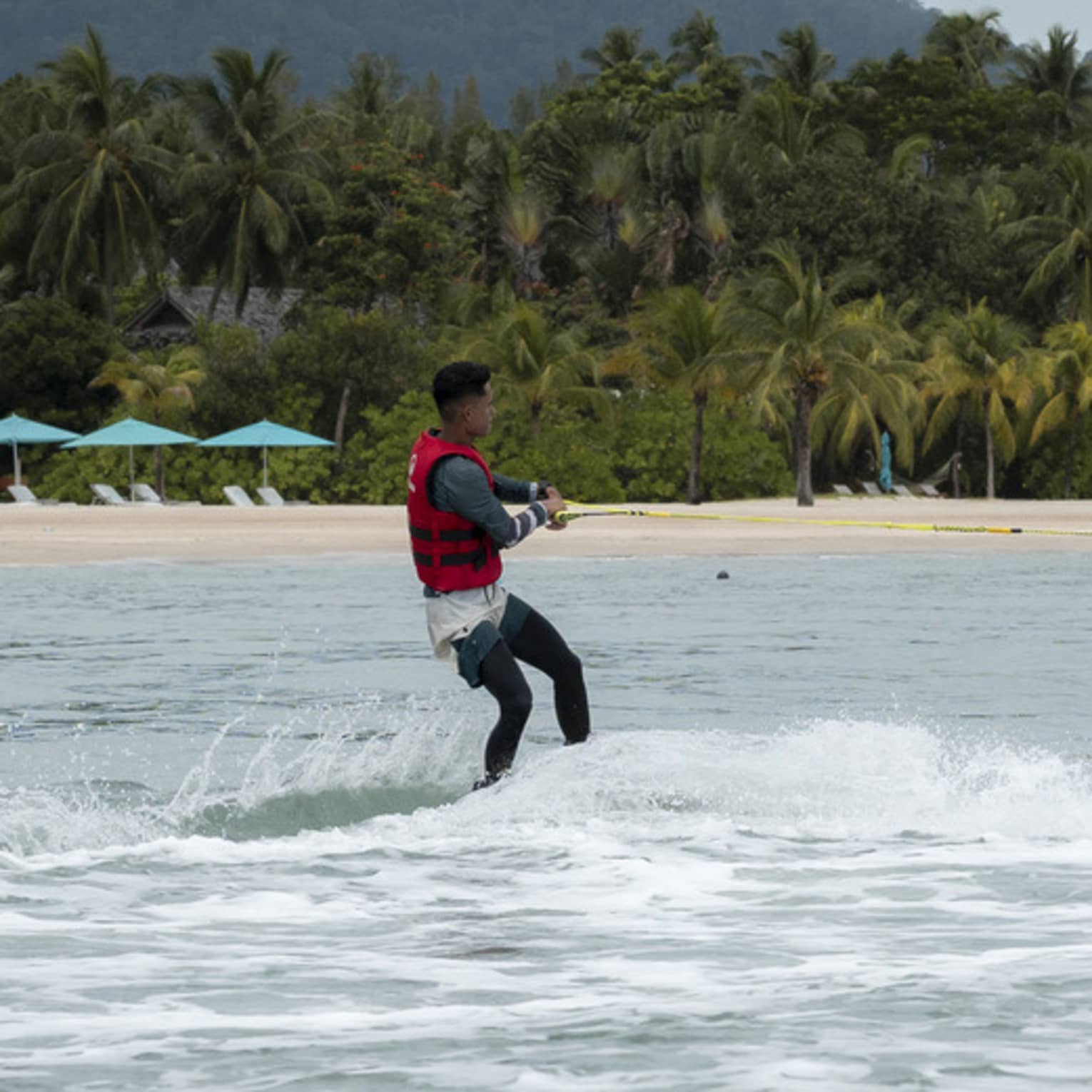 Person in red life jacket glides along the water on a water ski, the shore and copse of palm trees in the distance