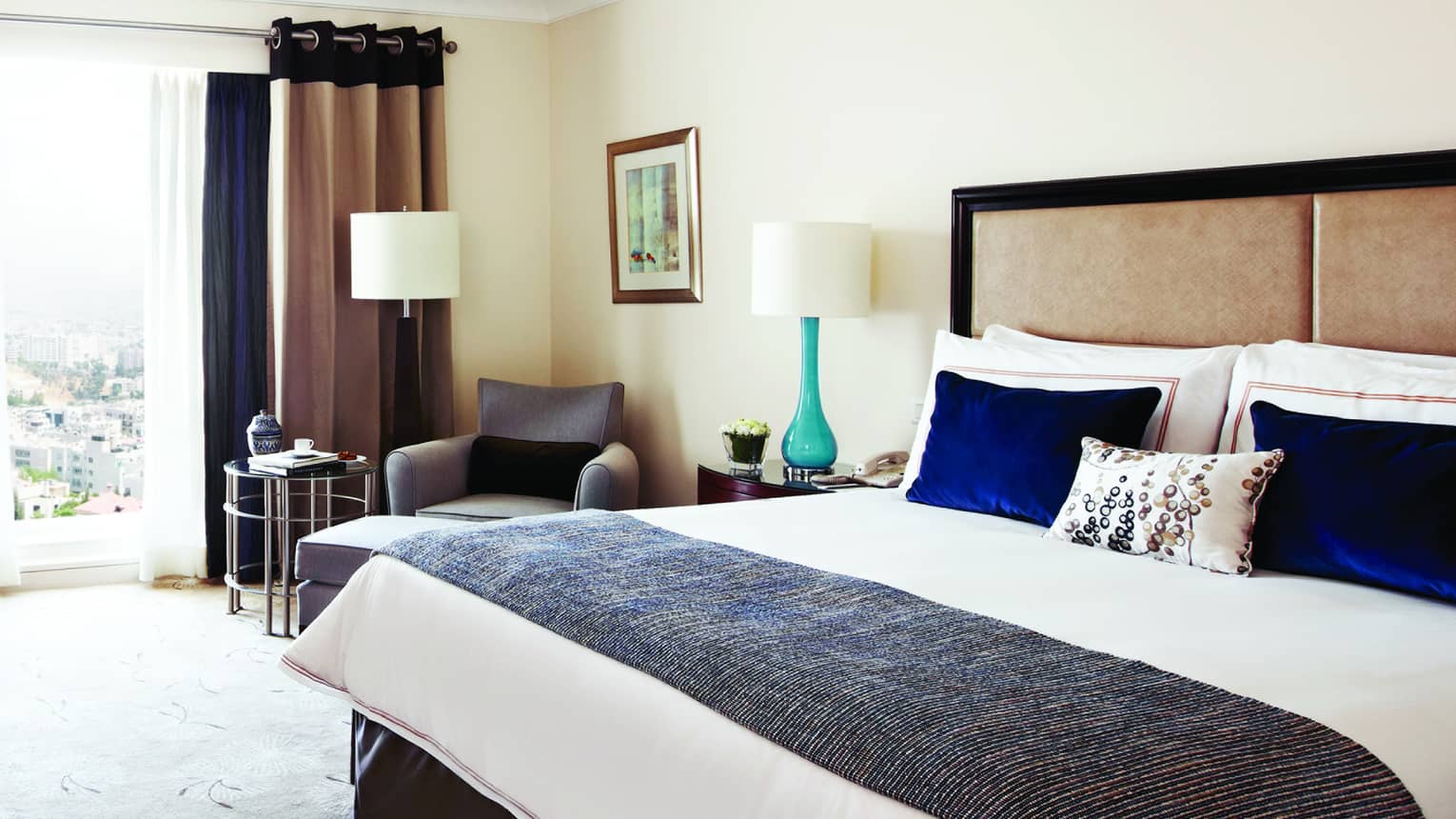 Superior room bed with blue velvet pillows, knit blanket, beige padded headboard, teal lamp, sunny window