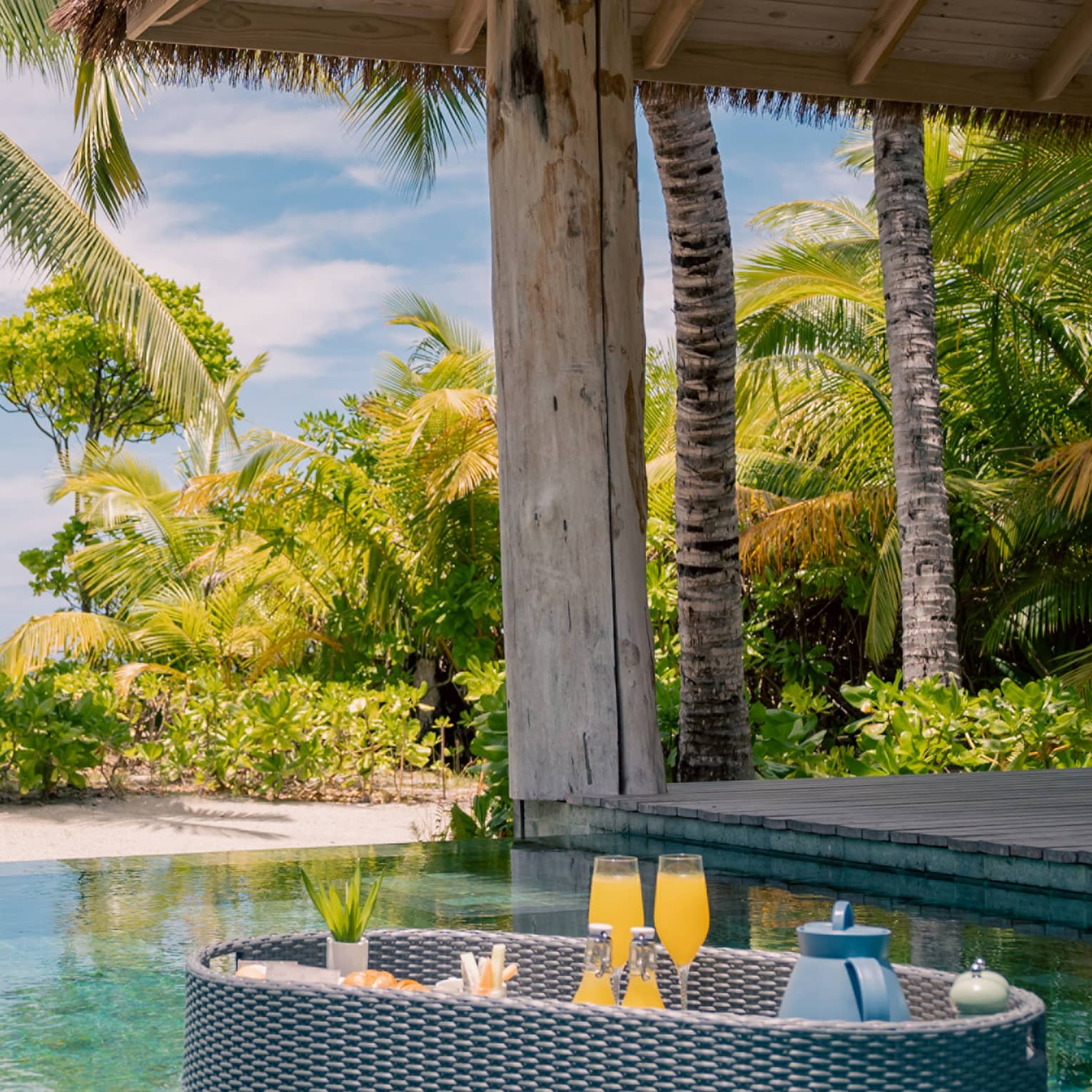 Wicker tray floats in a pool carrying two flute glasses with a bright orange drink, beside a tropical cabana and palm trees.
