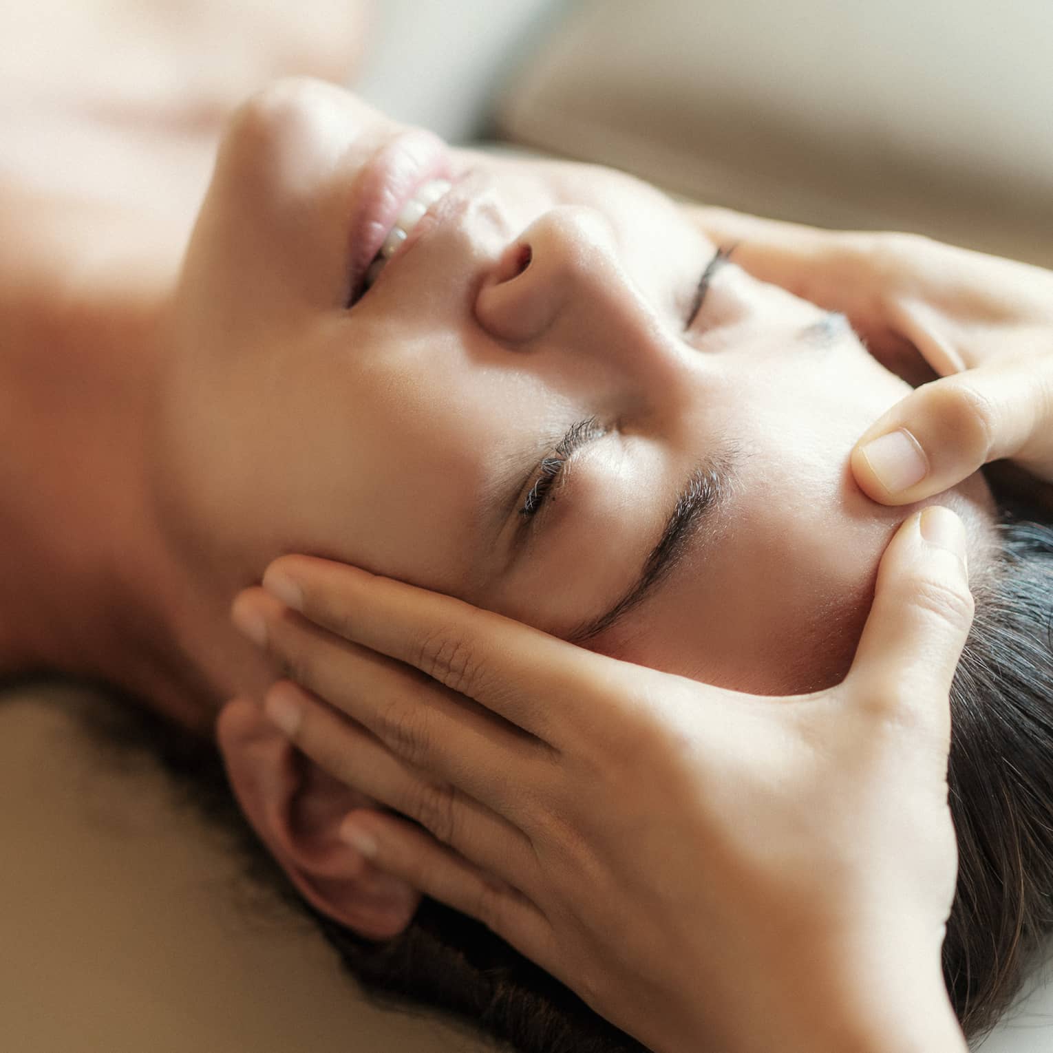 Spa facial, two hands rest on woman's forehead as she closes her eyes, lays under sheet on treatment table
