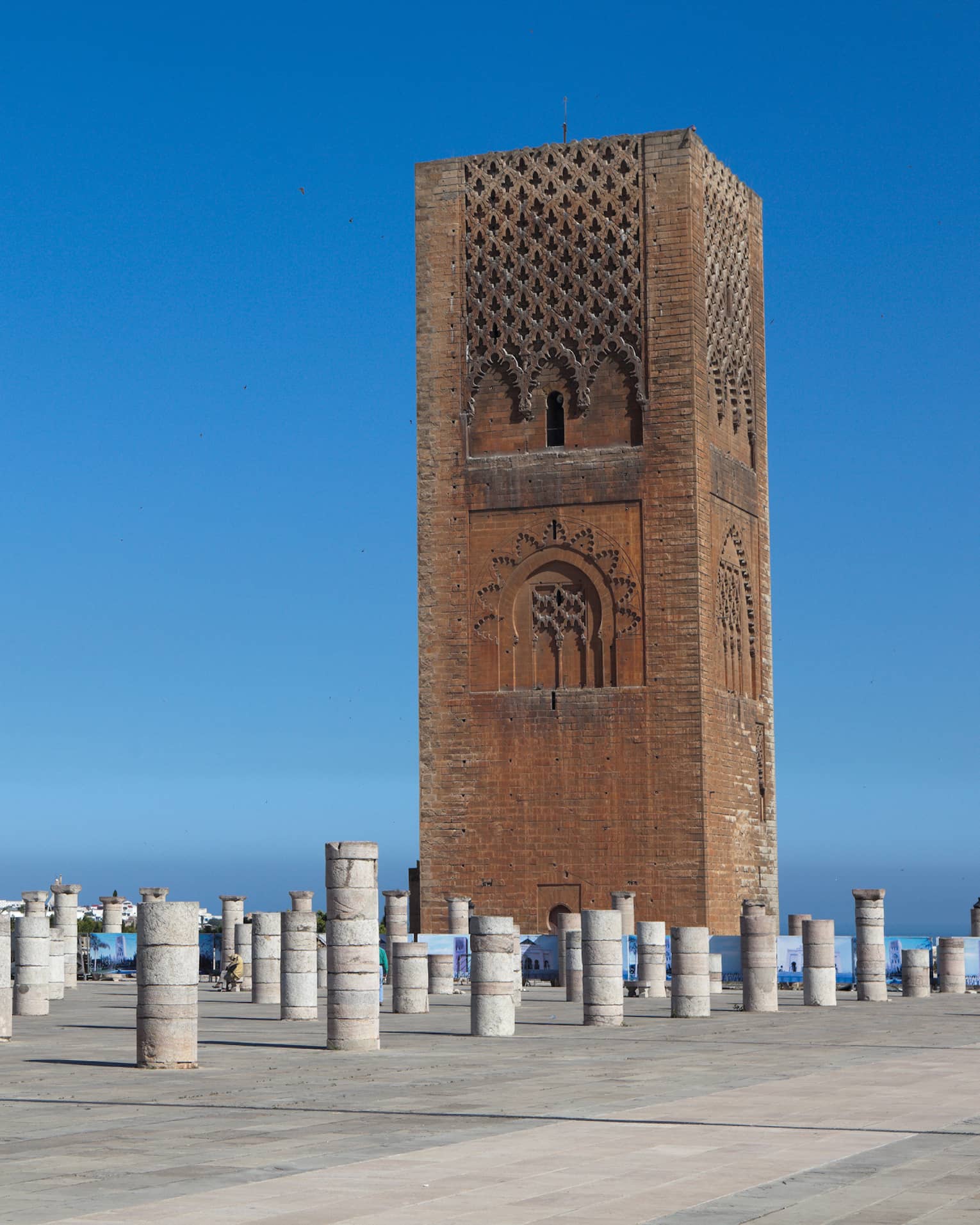 Against a blue sky, a tall rectangular red sandstone tower with decorative panels looms over dozens of short stone pillars.