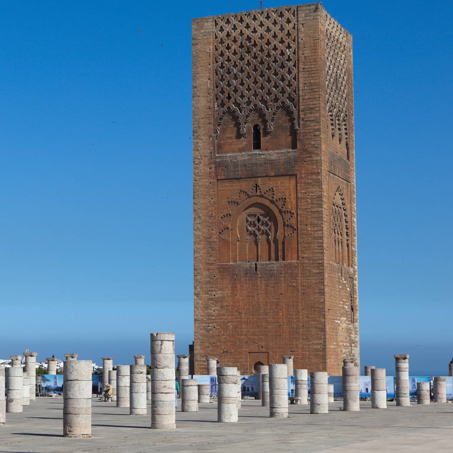Against a blue sky, a tall rectangular red sandstone tower with decorative panels looms over dozens of short stone pillars.