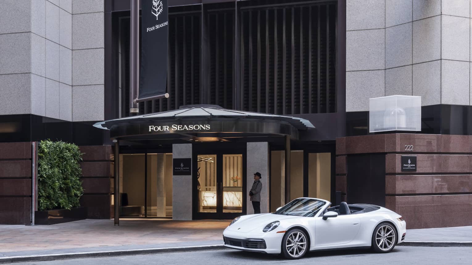 A white luxury sports caris parked in front of the massive Four Seasons hotel entrance, which is clad in pale grey granite.