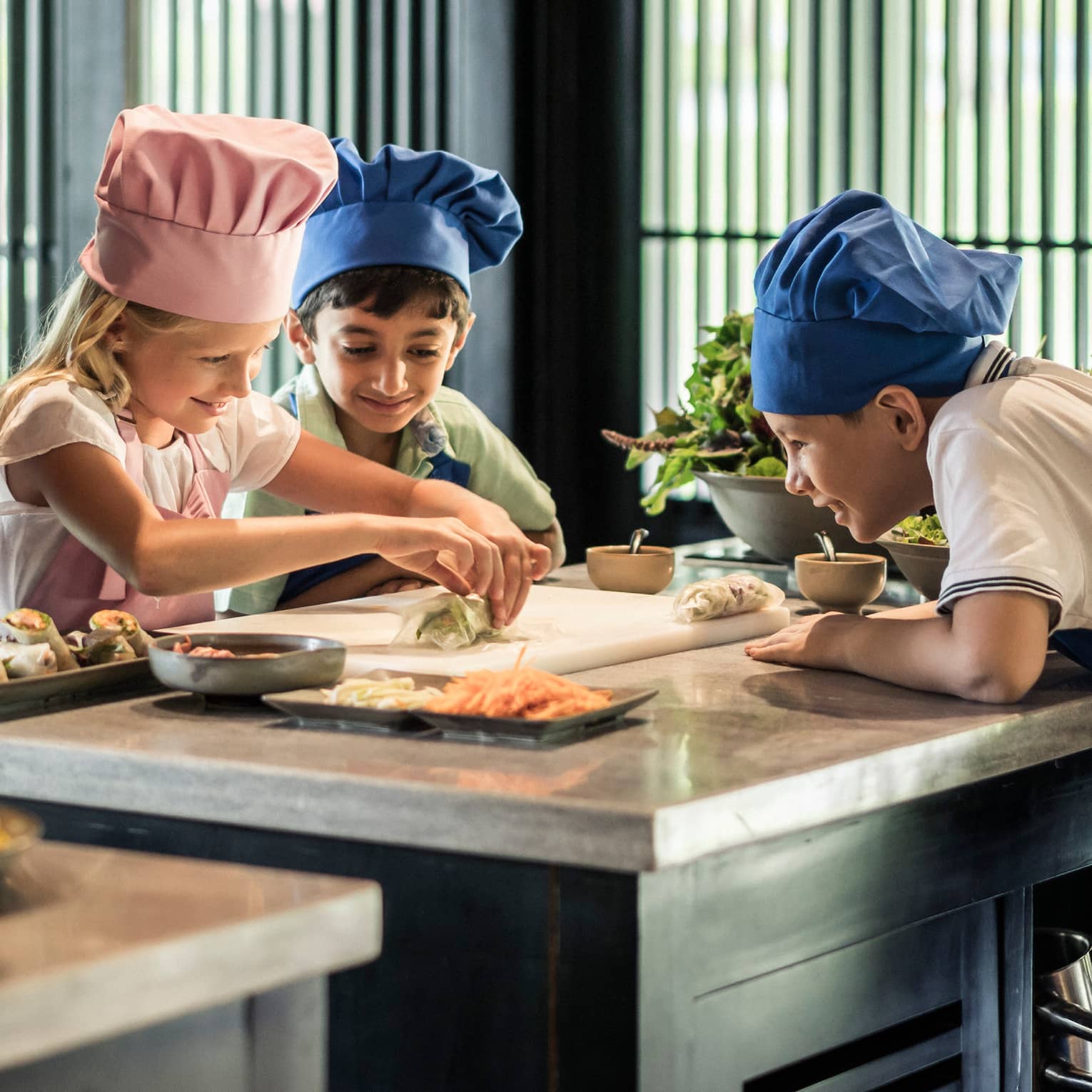 Chef watches as children wearing tall chef hats make spring rolls at counter