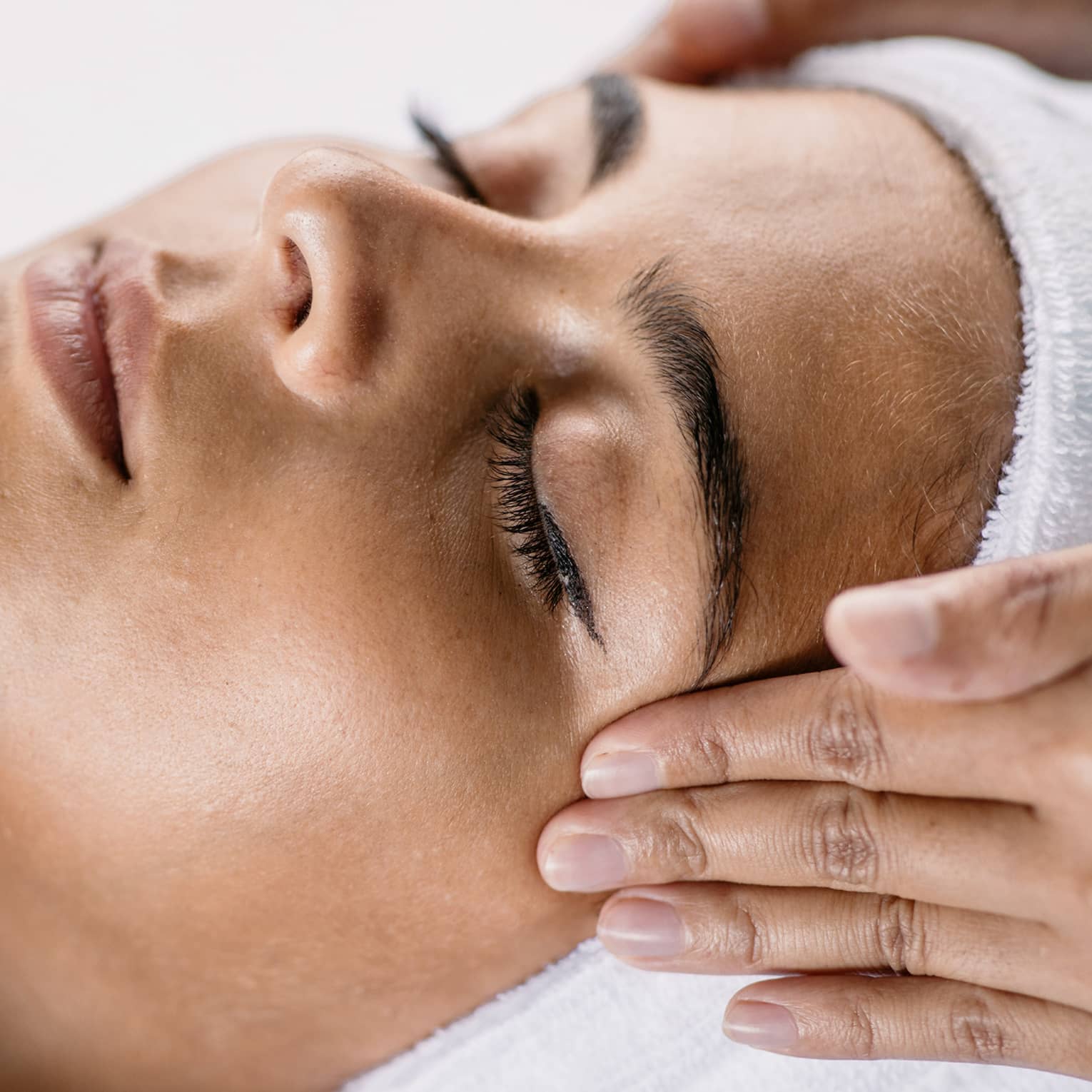 Hands massage woman's temples as she closes her eyes, spa towel around hair