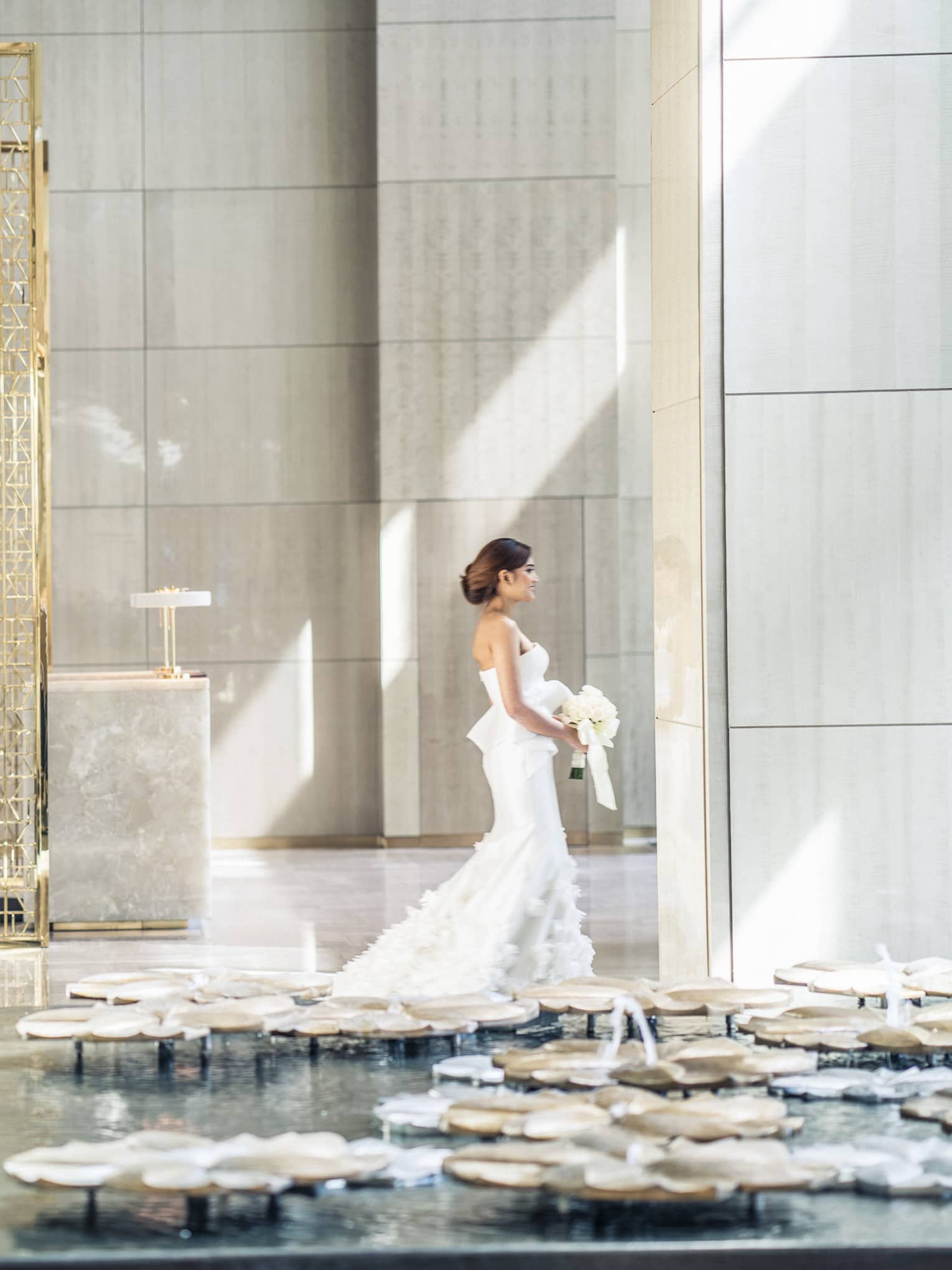 Sun rays illuminates a bride as she walks through a white walled lobby carrying her bouquet of white flowers