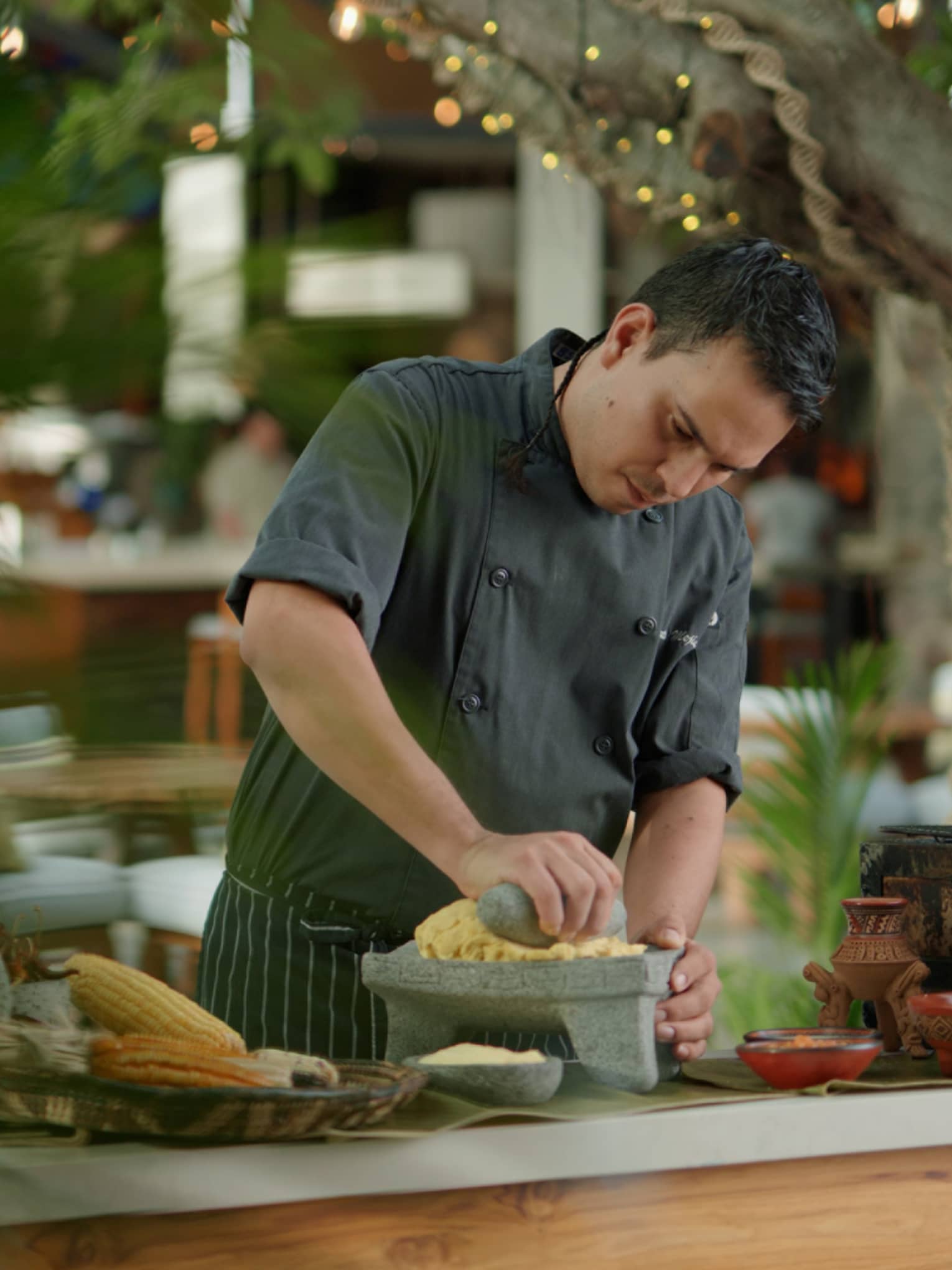 Chef wearing dark grey uniform mashes corn using a stone mortar and pestle