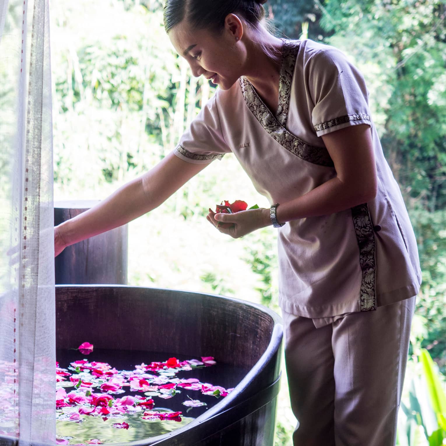 Hotel staff drops colourful flower petals in free-standing copper bathtub draped with sheer white net 
