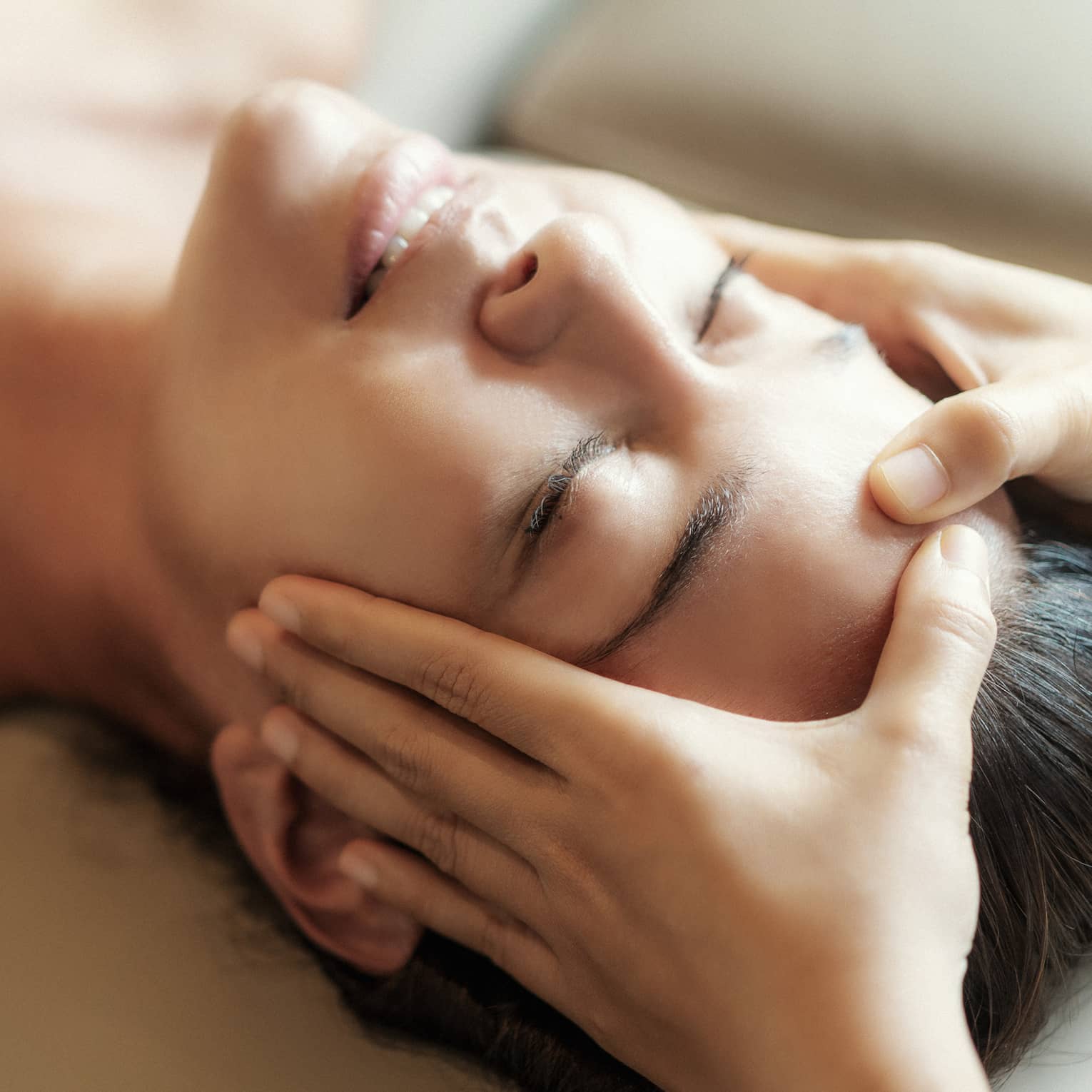 Spa facial, two hands rest on woman's forehead as she closes her eyes, lays under sheet on treatment table