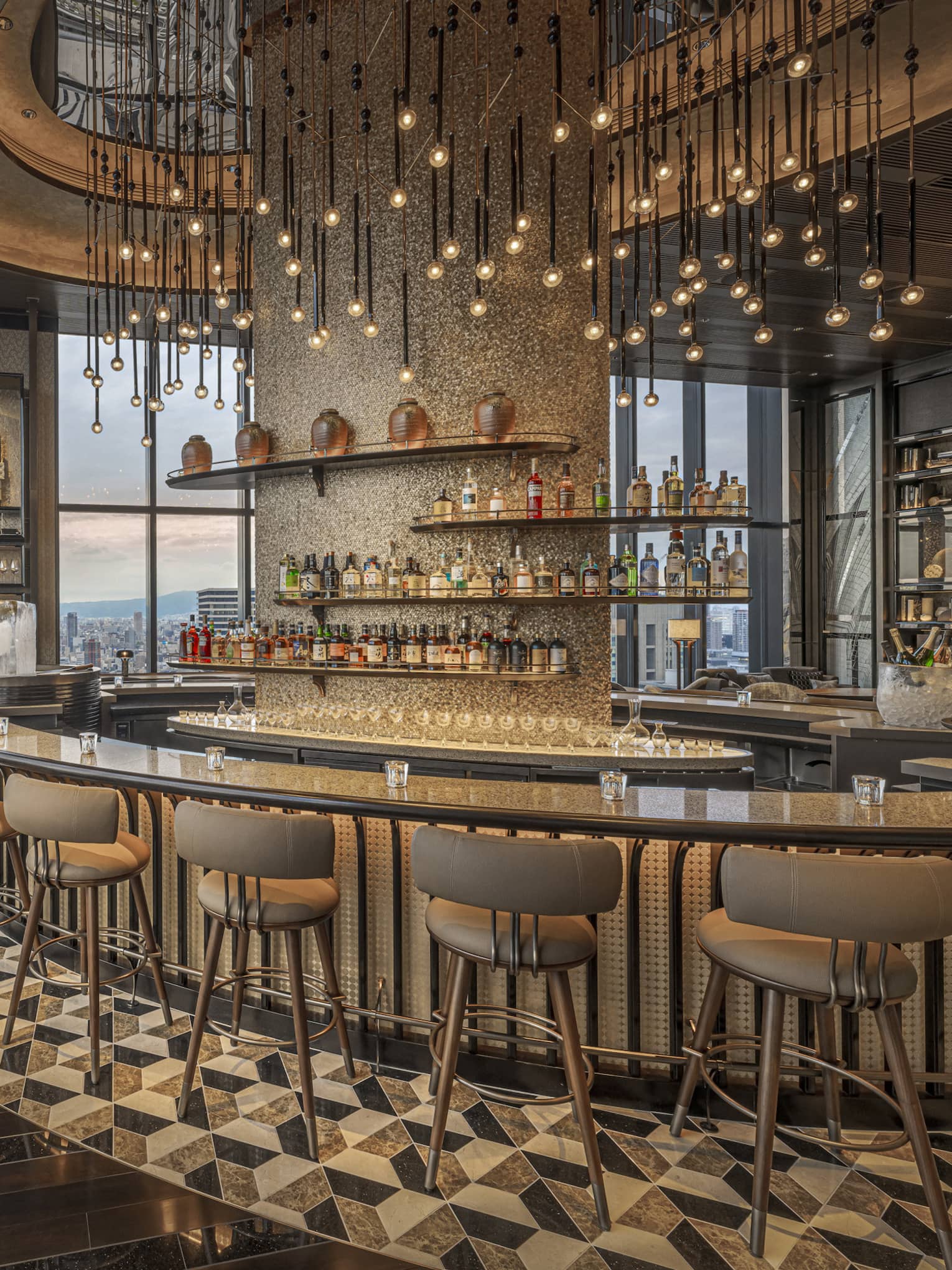 Large circular granite-topped bar in a modern luxury restaurant surrounded by a bank of windows overlooking city at dusk.