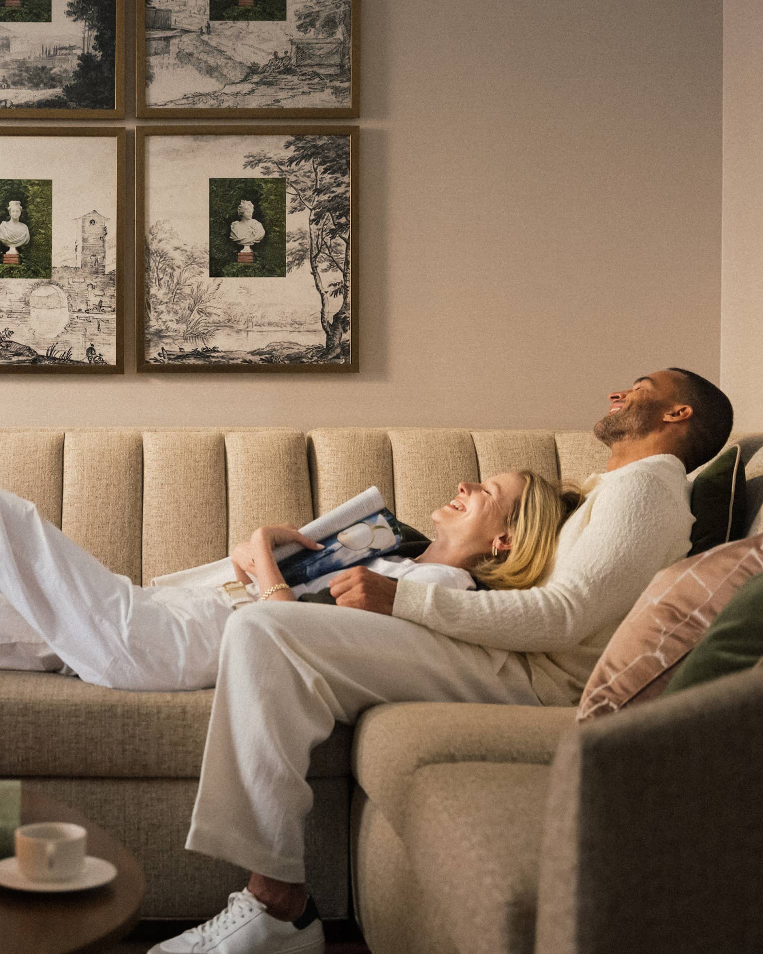 Two guests laying on the couch in a guest room with picture frames on the wall and a bouquet of flowers on the coffee table
