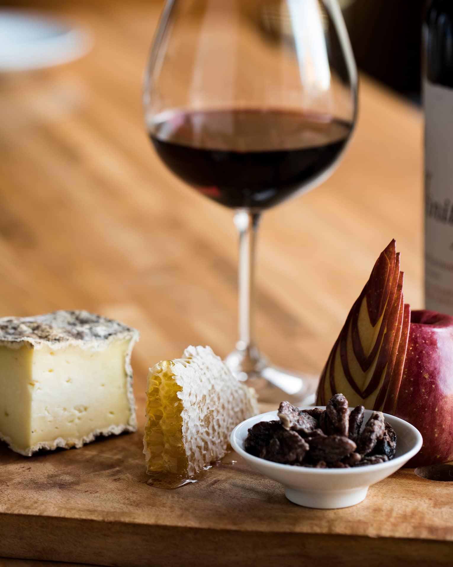 A glass of red wine with a luxurious wooden tray holding cheese, honeycomb, honey, sliced apple, nuts and crackers.
