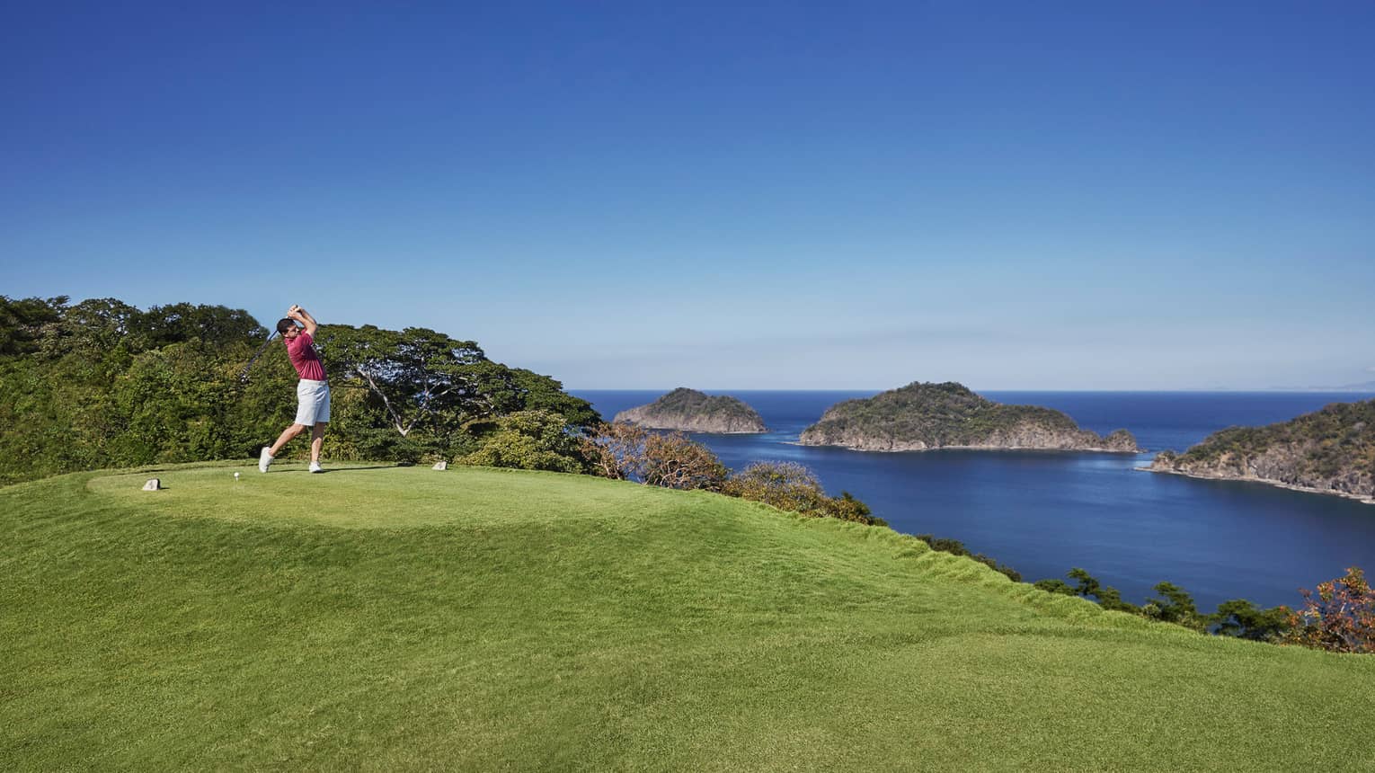 Man swings golf club on course green high above ocean