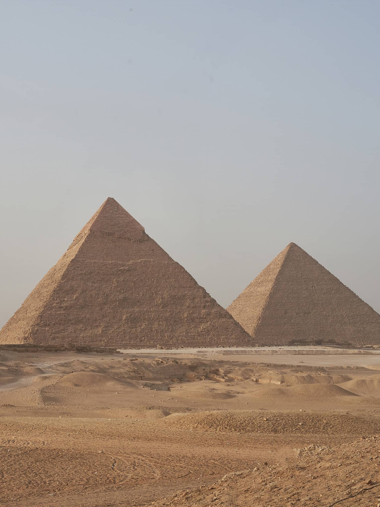 In the arid desert, a line of three towering pyramids dwarf two smaller pyramids before them, under a hazy blue sky. 
