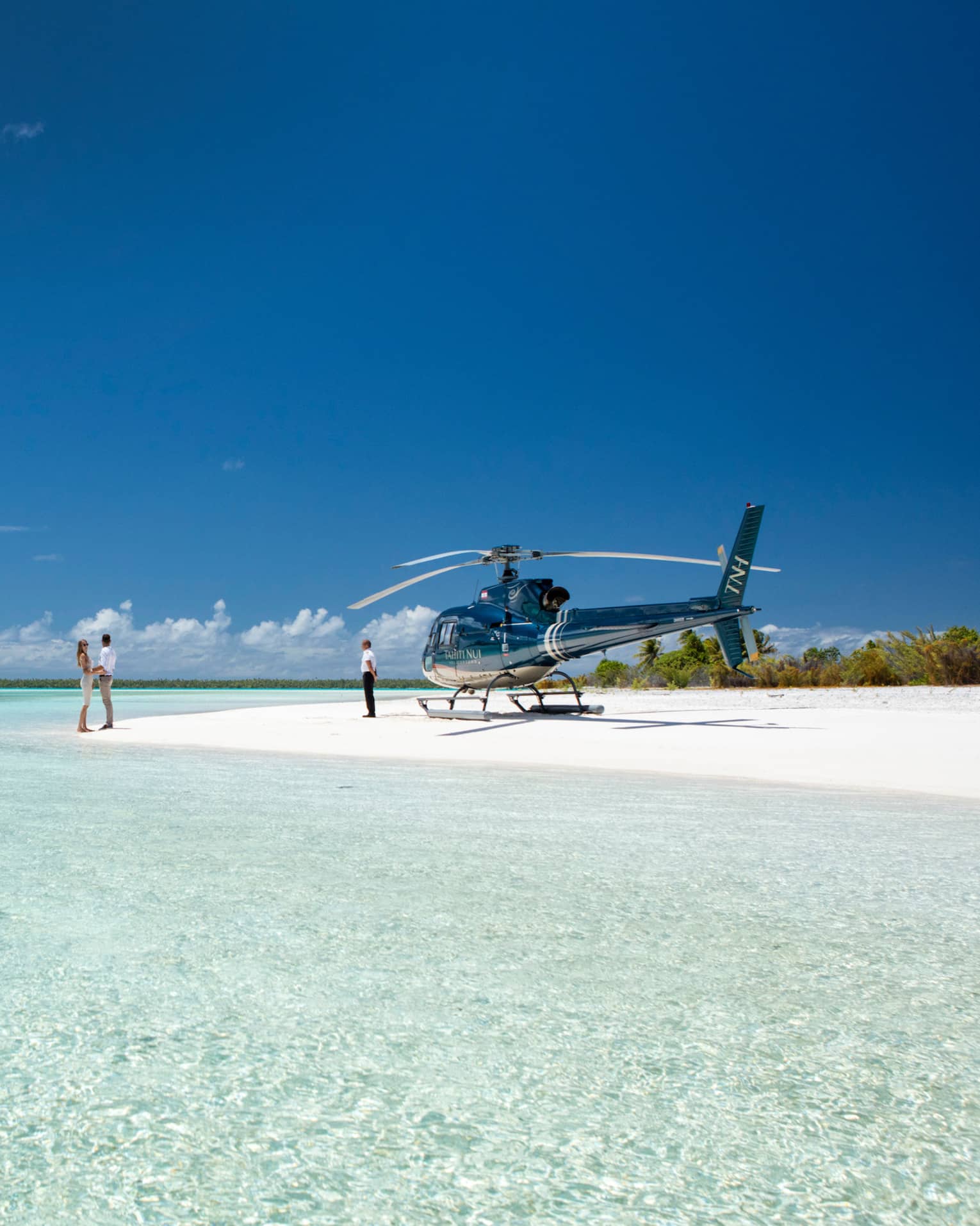 A couple holds hands on a white sand beach beside the pastel blue ocean; a parked helicopter and its pilot wait nearby.