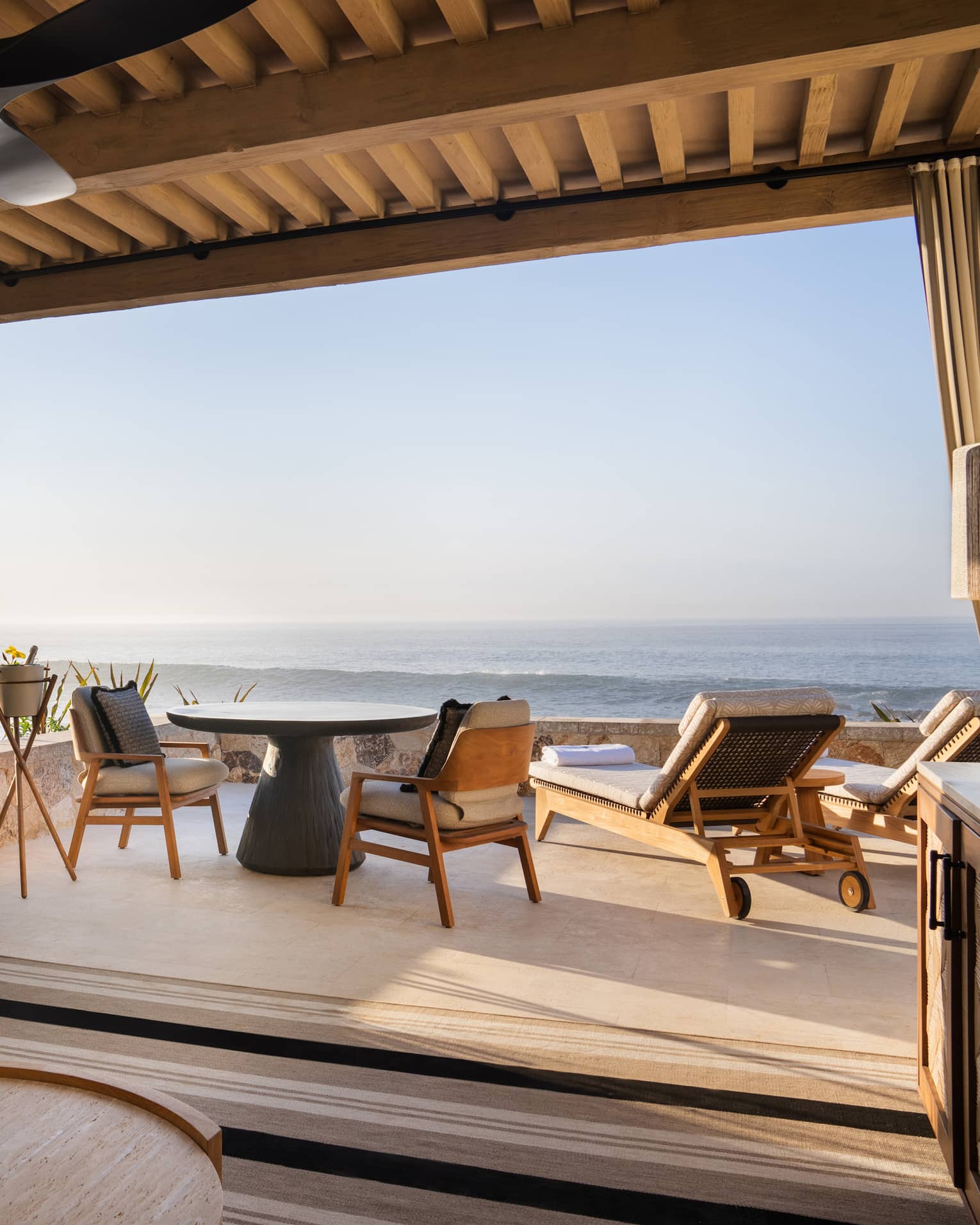 Oceanfront cabana with beige sofa, lamp and round wood coffee table under a roof with a round table and chairs and two lounge chairs out on the open-air patio overlooking the ocean