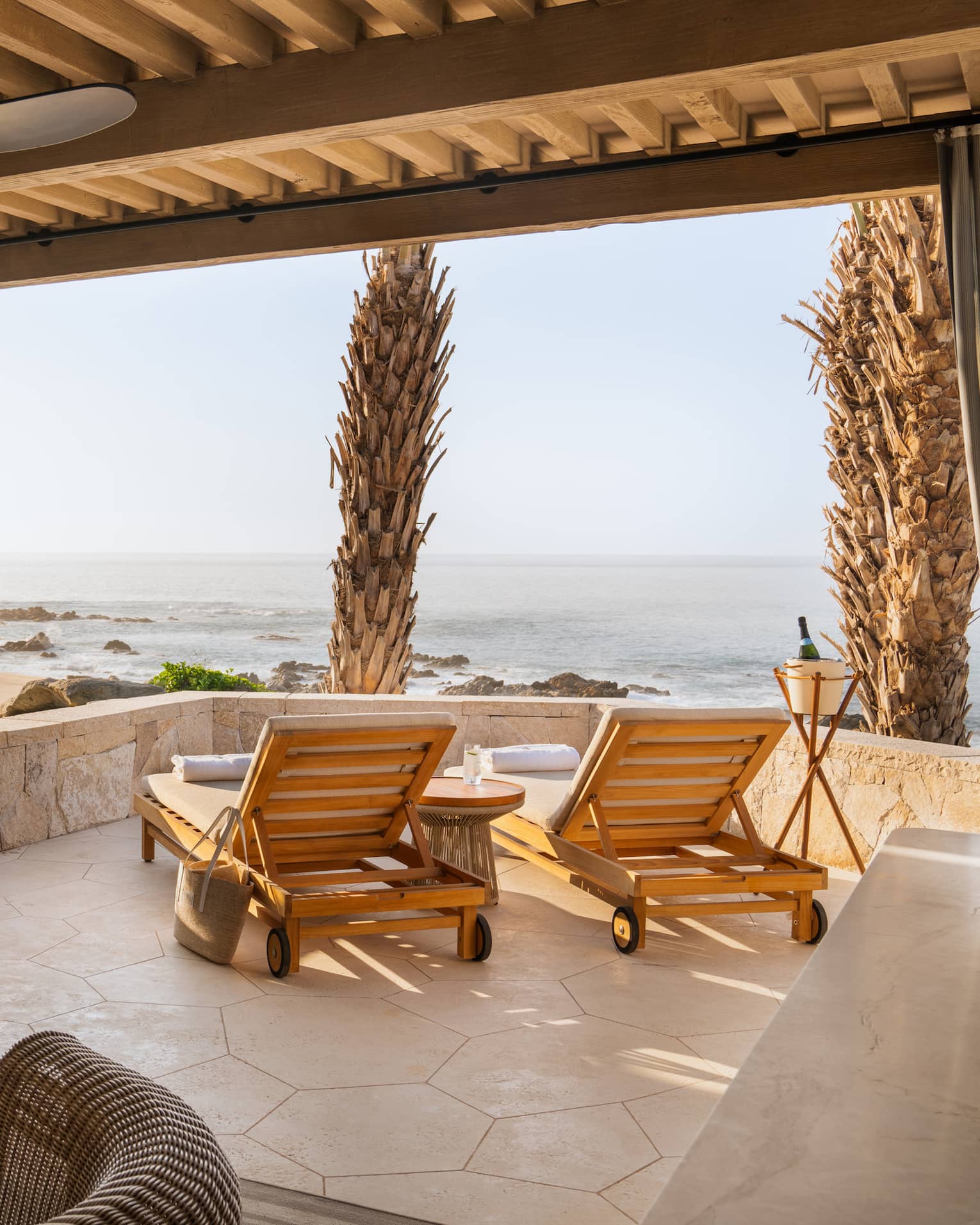View from inside a neutral cabana with lounge chairs by a low stone wall looking out towards a rocky ocean shoreline.