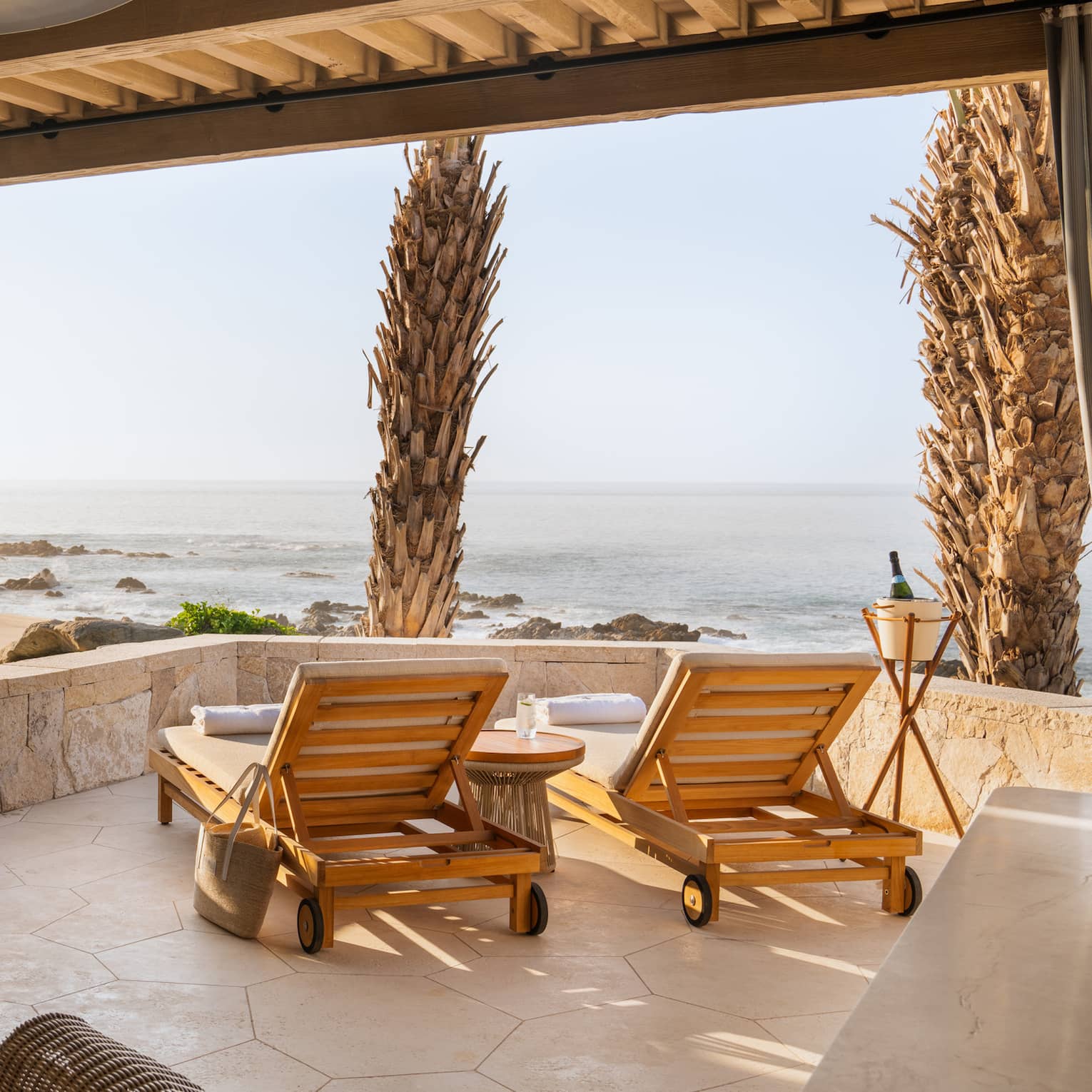 View from inside a neutral cabana with lounge chairs by a low stone wall looking out towards a rocky ocean shoreline.