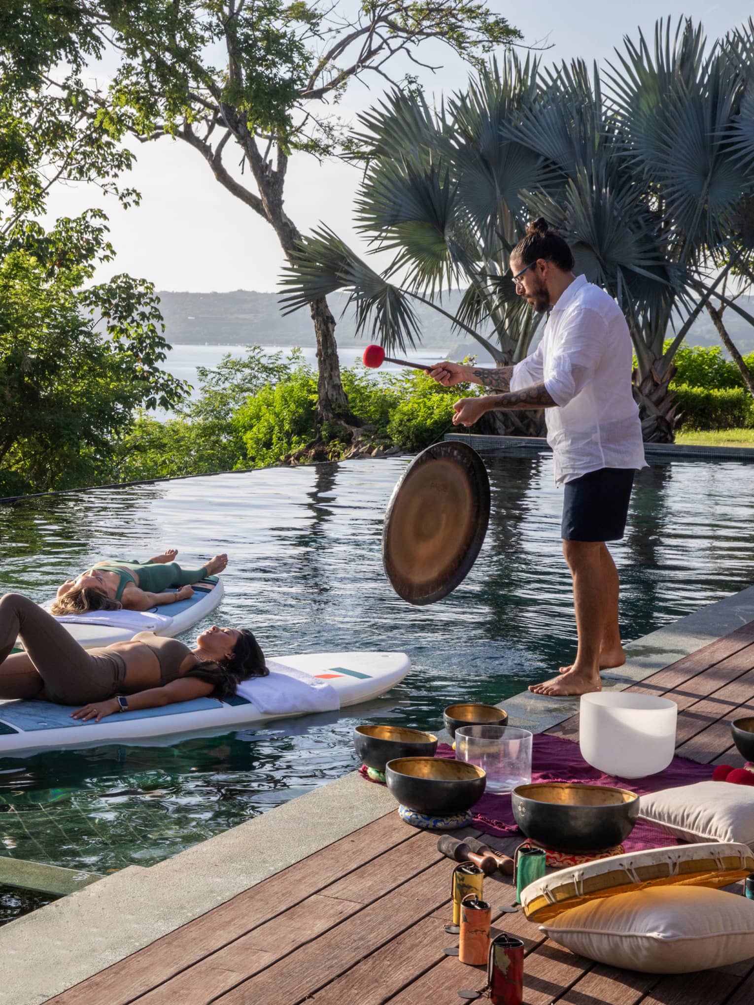 Three people float on stand-up paddleboards in a private pool while someone plays a gong on the deck