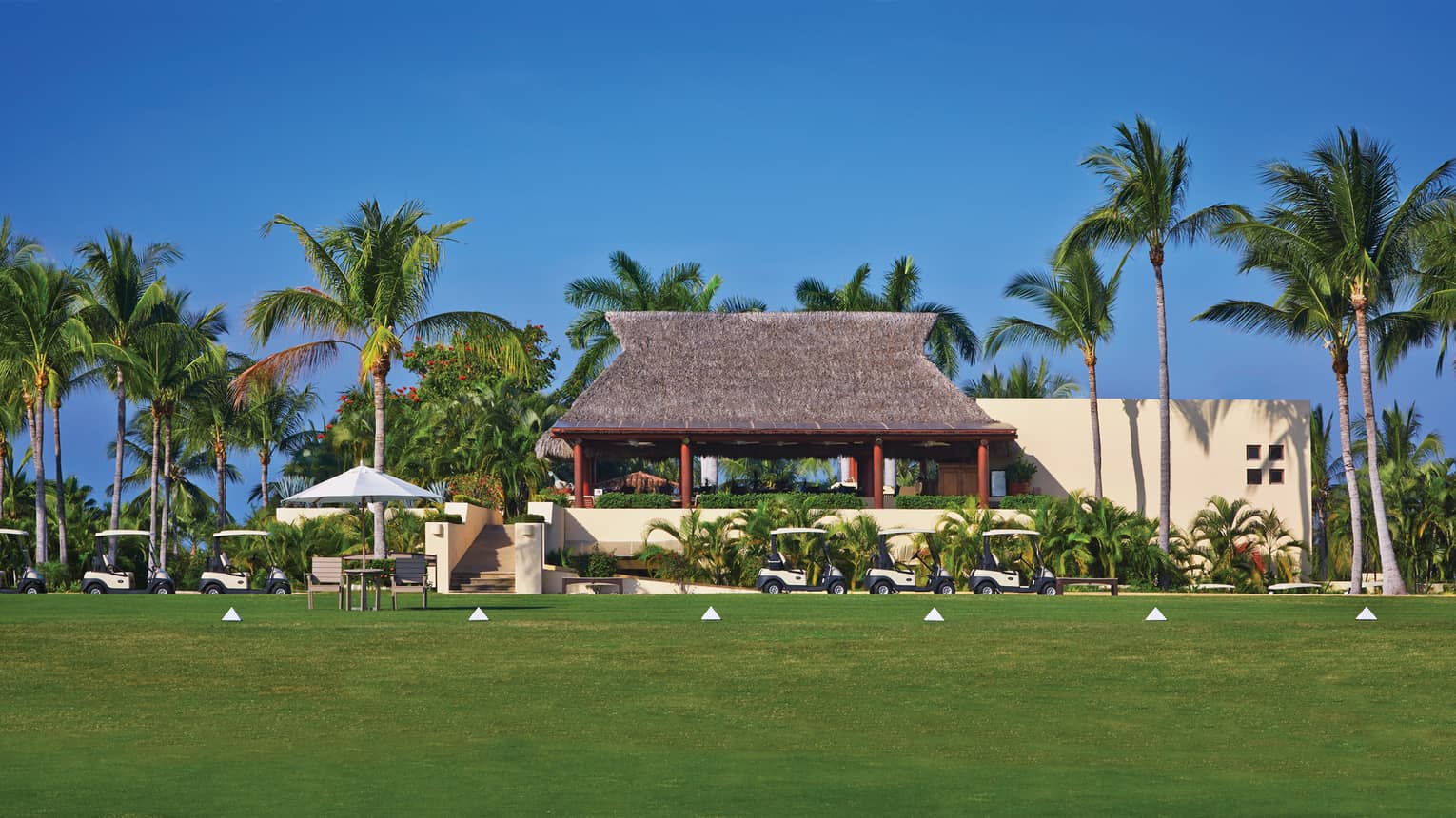 View across golf course green to tiki pavilion, palm trees and row of golf carts