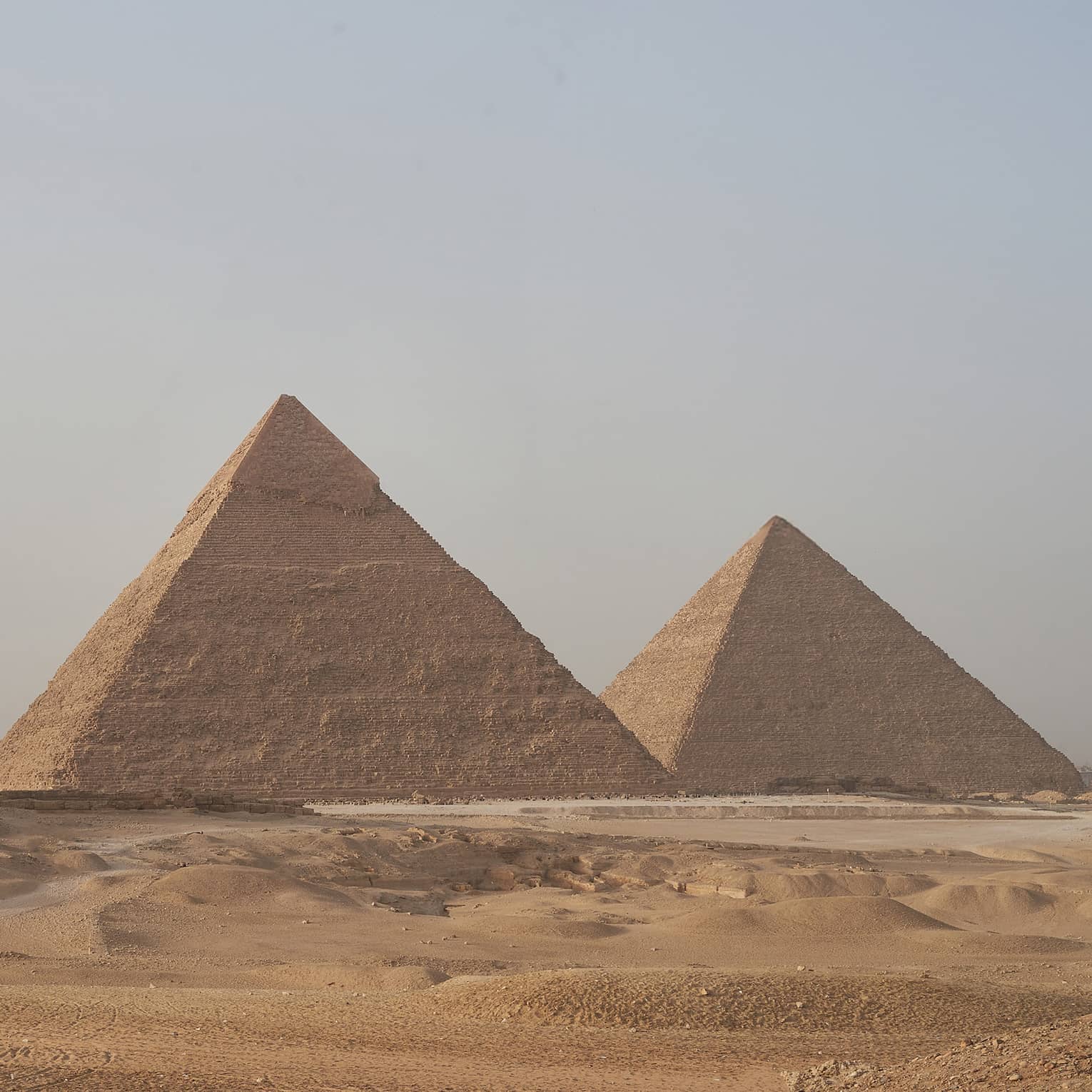 In the arid desert, a line of three towering pyramids dwarf two smaller pyramids before them, under a hazy blue sky. 