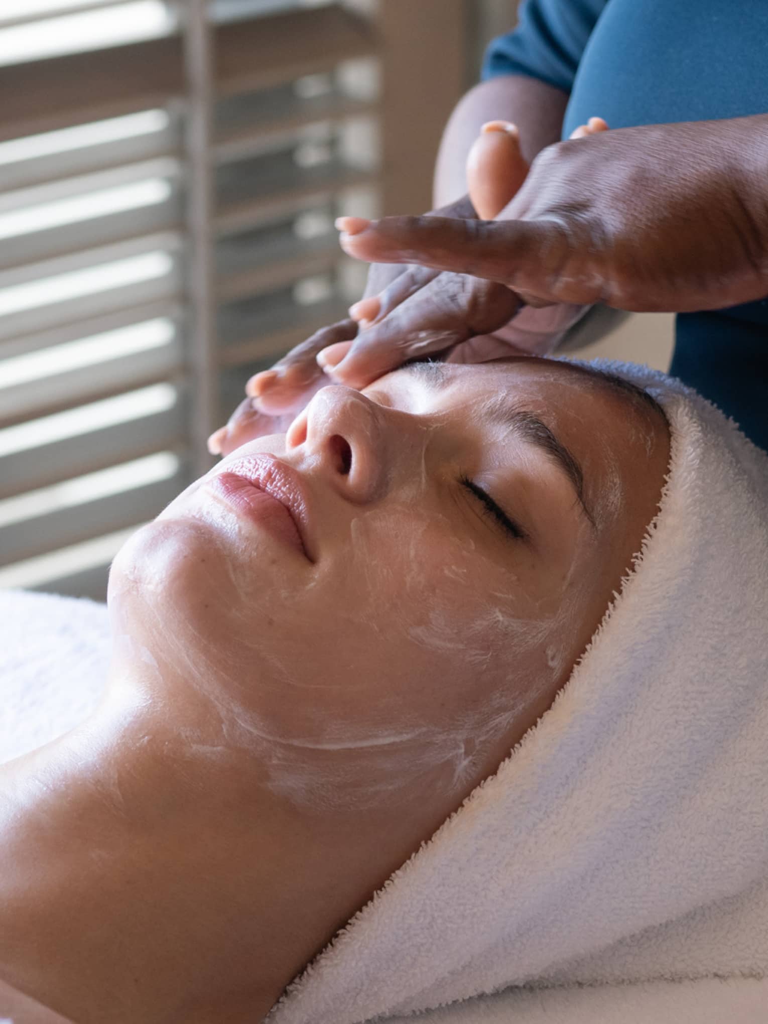 Woman gets facial. A white towel protects her hair as white cream is massaged into her face.
