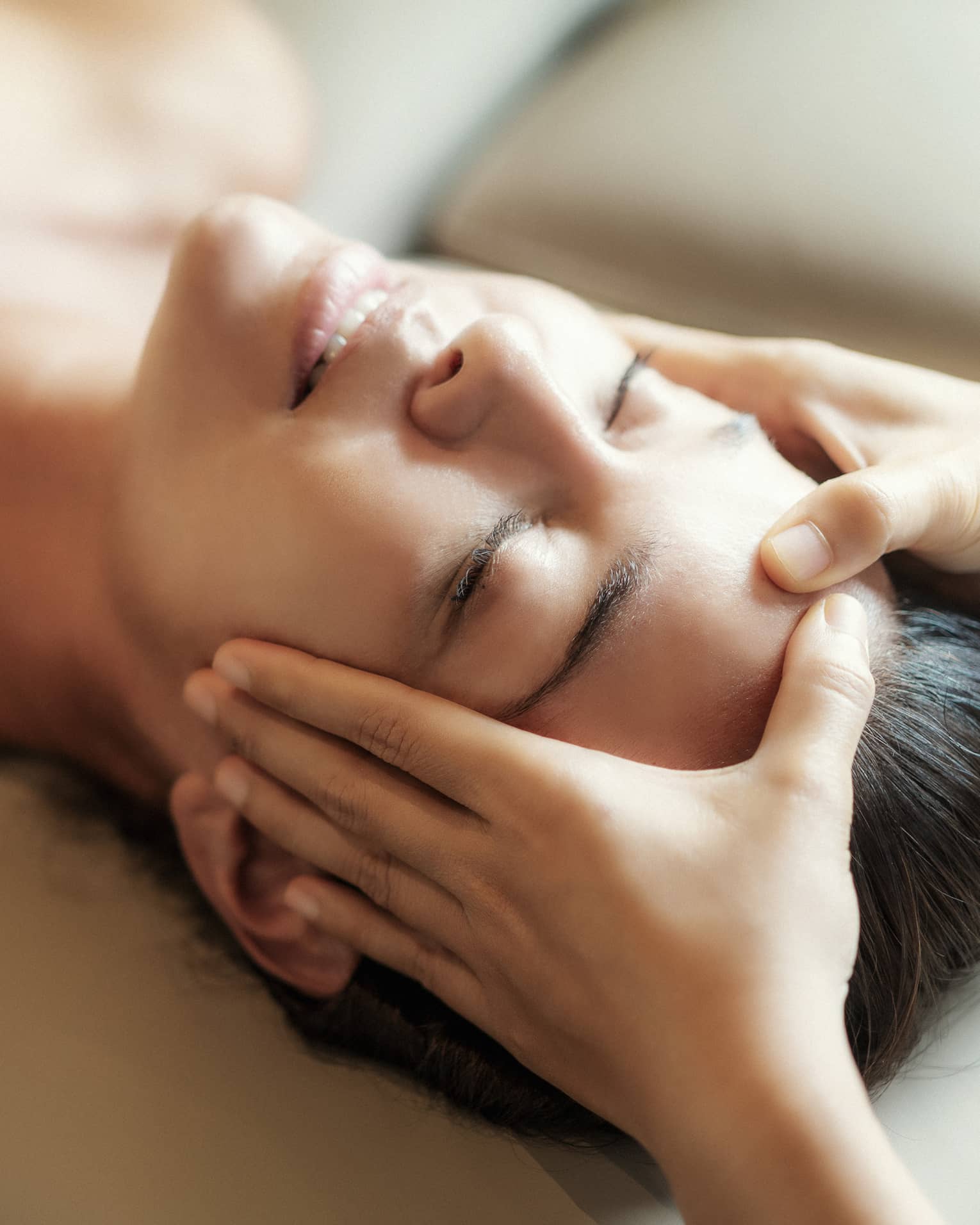 Spa facial, two hands rest on woman's forehead as she closes her eyes, lays under sheet on treatment table