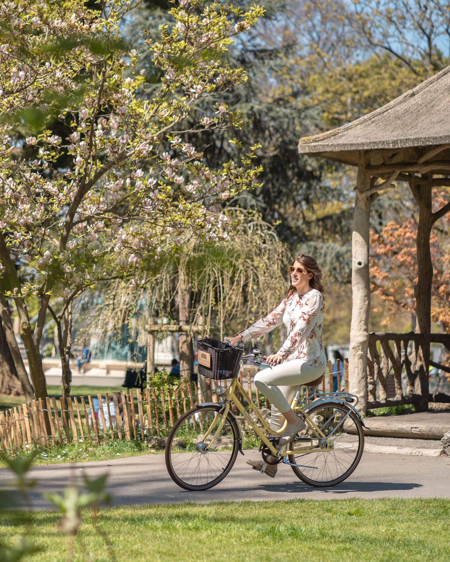 Side view of a smiling guest on an bike with a basket coasting past mixed forest and spring blossoms in a sunny park.