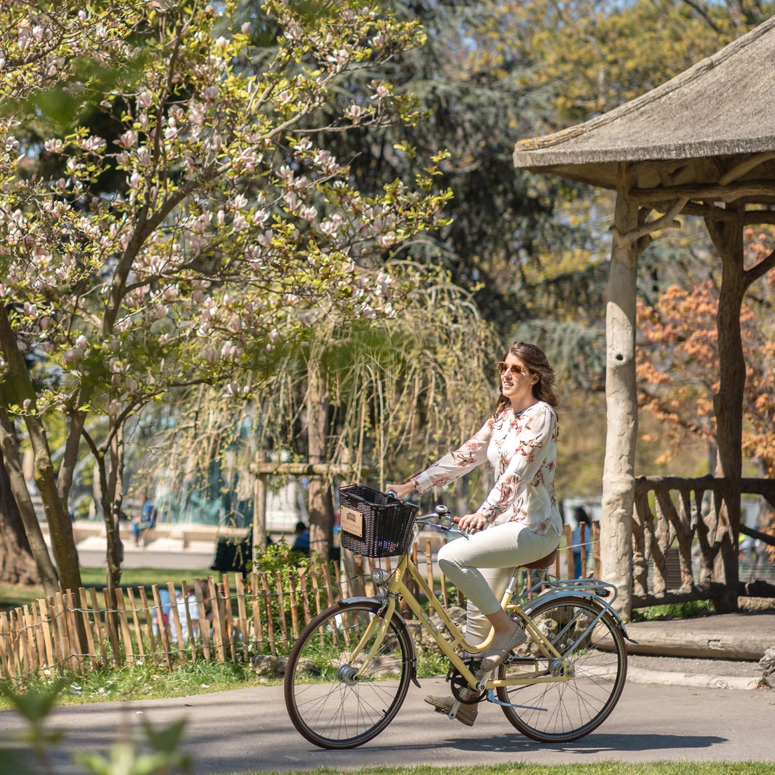 Side view of a smiling guest on an bike with a basket coasting past mixed forest and spring blossoms in a sunny park.