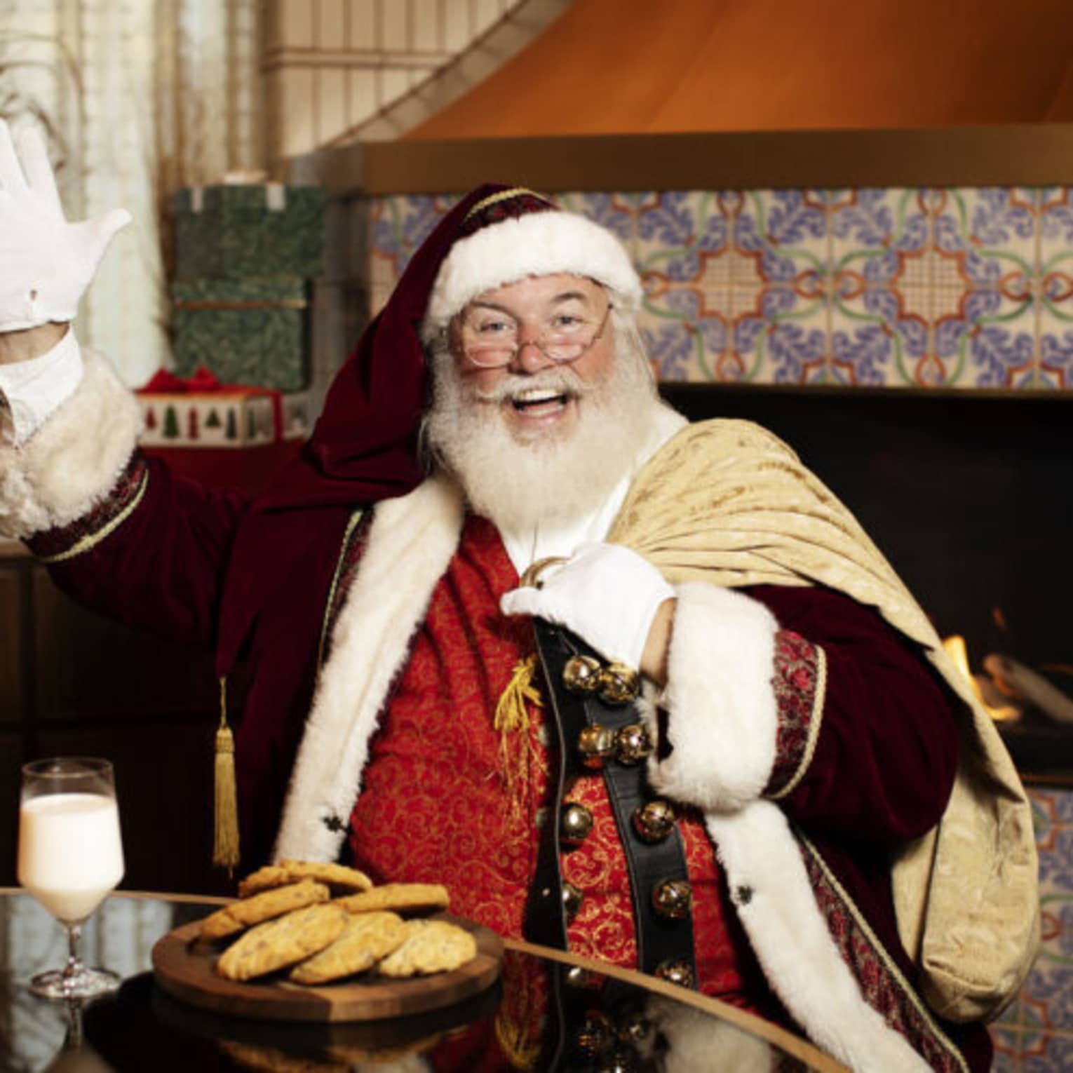 Santa Claus sitting at a table with a plate of cookies and glass of milk, and a fireplace in the background.