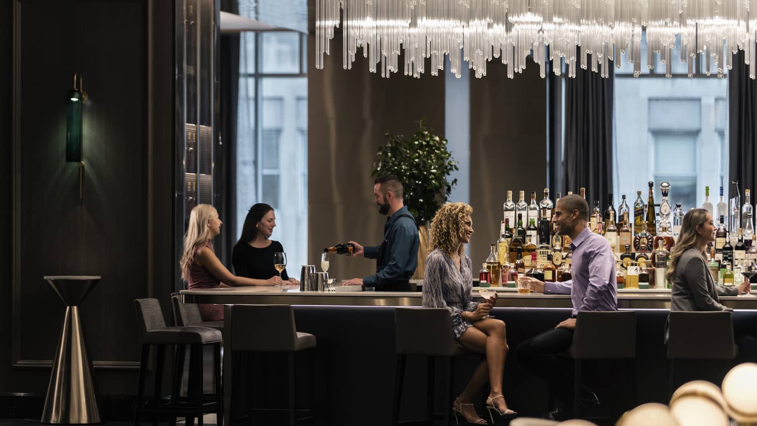 People seated at distance around dark modern bar and bartender under large chandelier