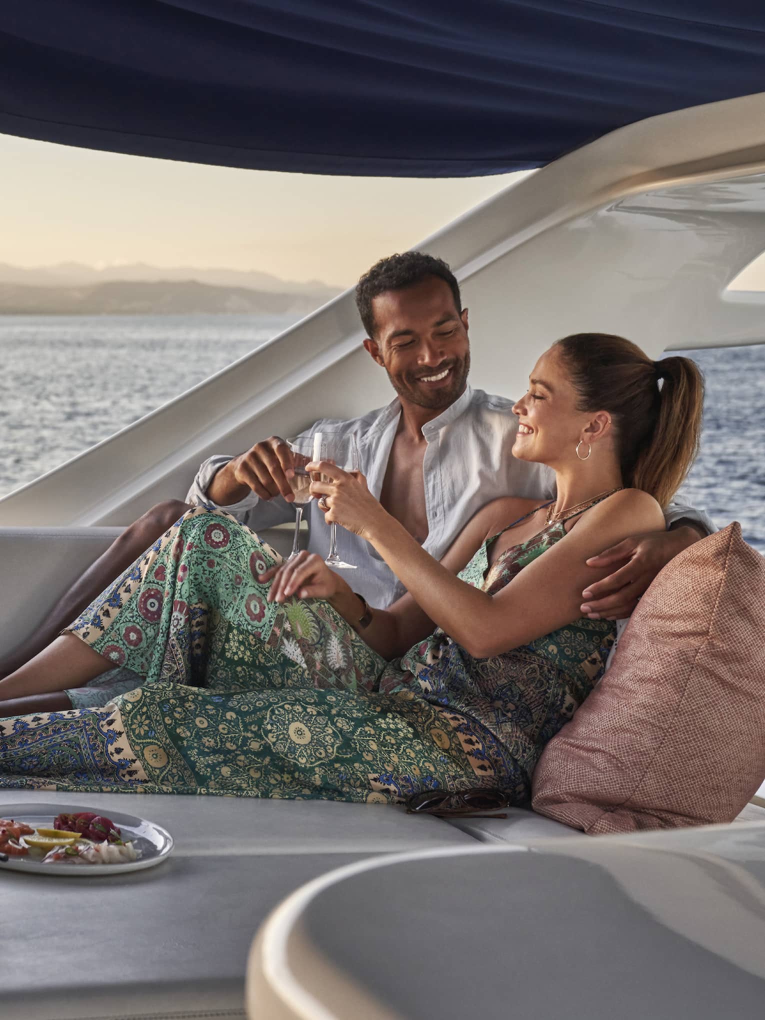 Aboard a boat in open air under a sunshade, a cuddling couple clink wine glasses amid cushions, dishes and an ice bucket.