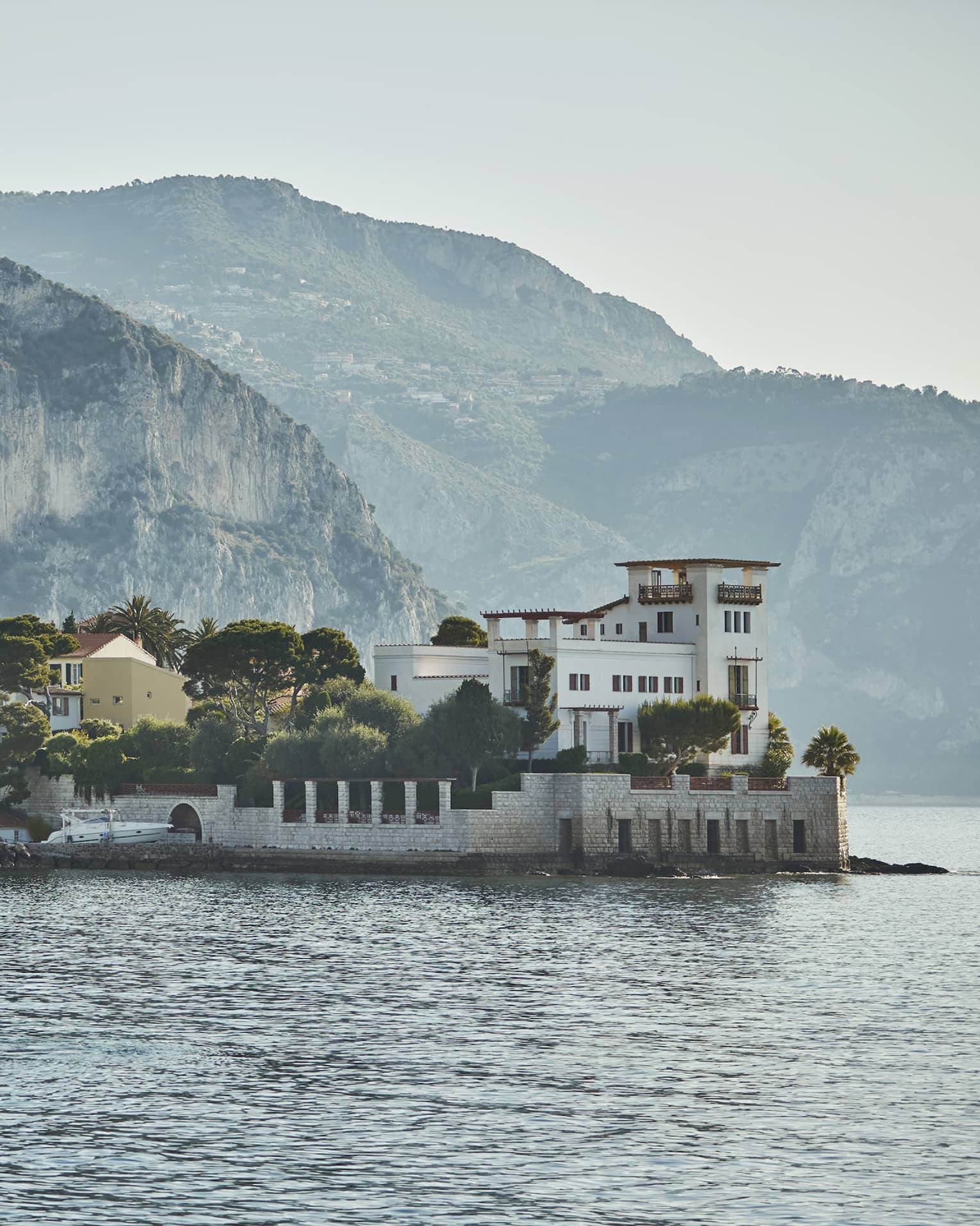 Cliffs and hills loom above a forested village along the water; a speed boat is anchored in front of a stone breakwater.