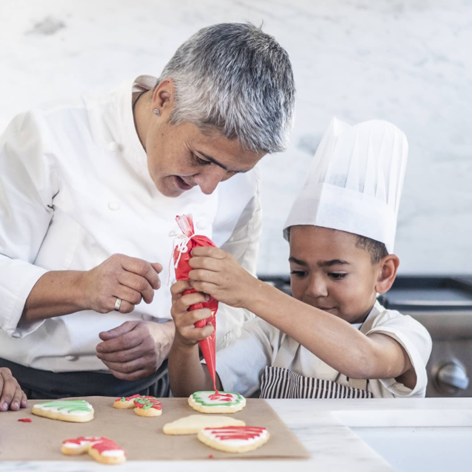 Two children in chef’s hats and aprons in a stark, white kitchen; a chef watches closely as one pipes icing onto a cookie.