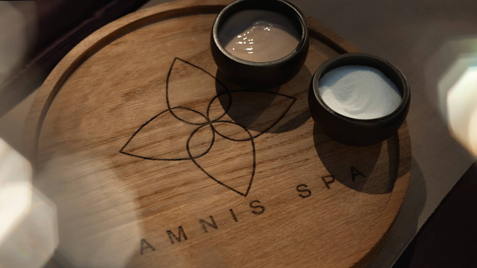Wood tray with Amnis Spa logo and two small black bowls with cream and salt