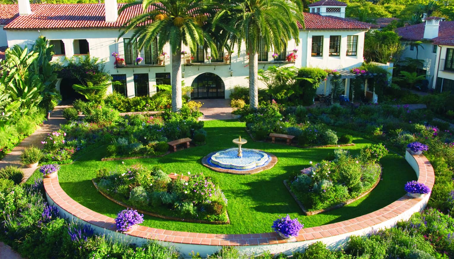 Lush green courtyard with a fountain in the center, flanked by two large palm trees and a Spanish Colonial-style building in the background, overlooking the mountains