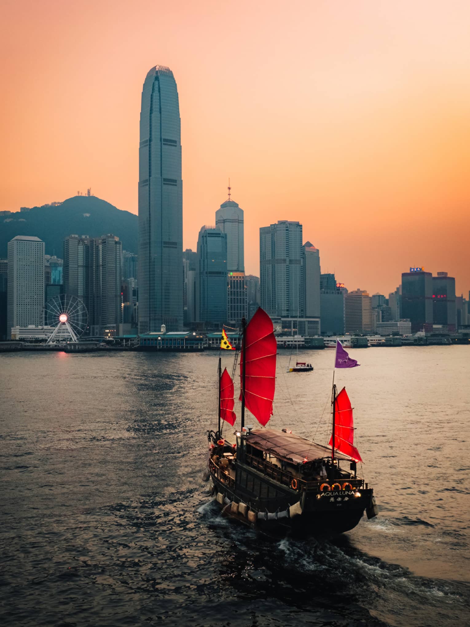 Boat with red sails on harbour in Hong Kong at sunset with city buildings at the shore