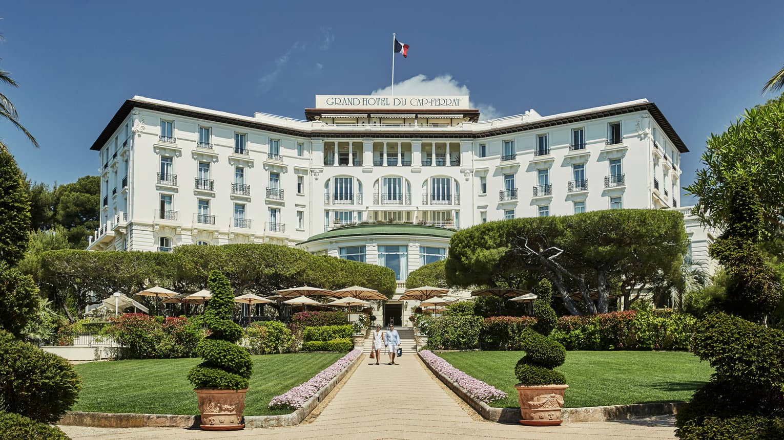 Two people walk down sunny stone path under large Grand-Hotel du Cap-Ferrat