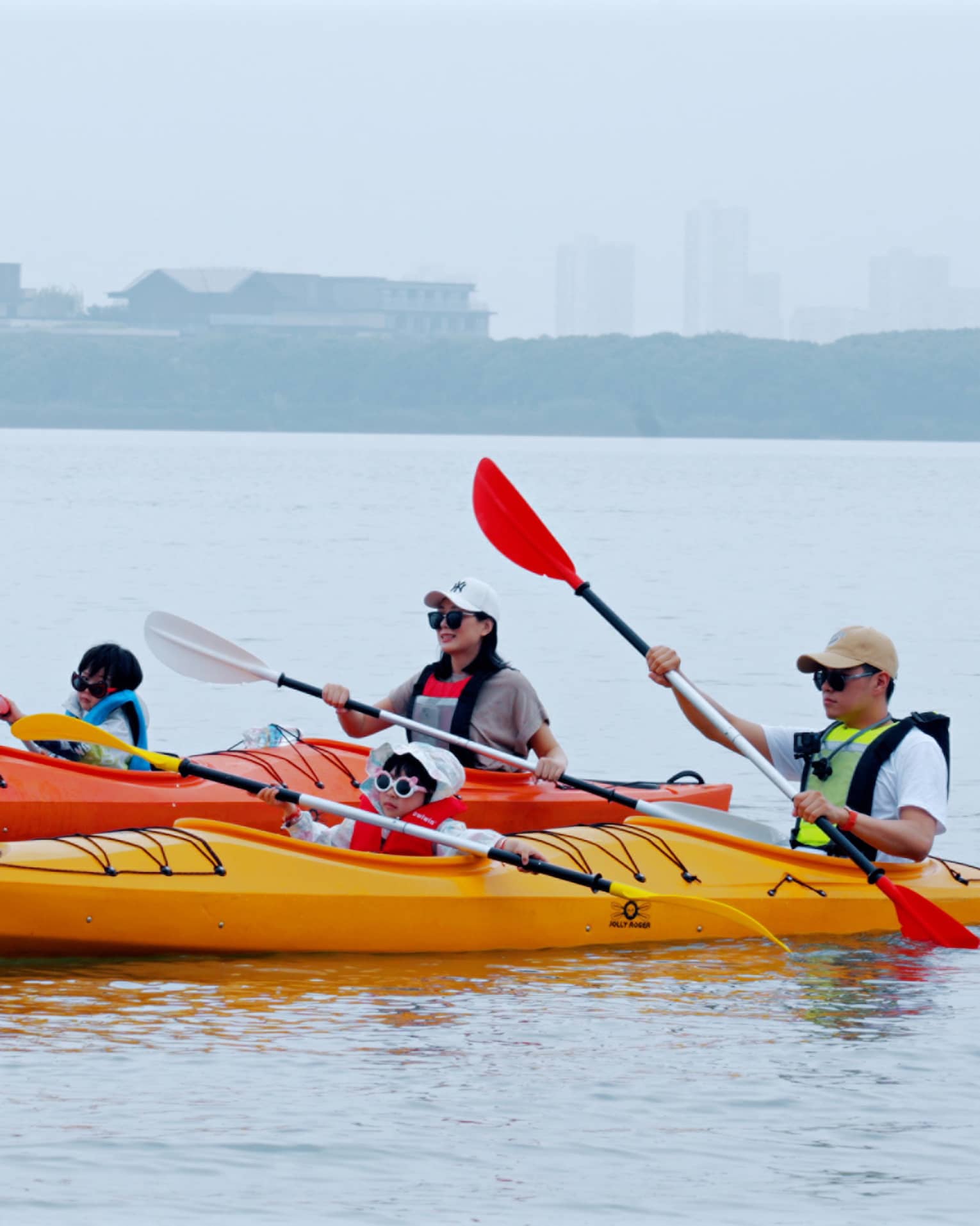 A family of four glides along serene waters, two people per kayak, distant terrain and buildings blurred by misty fog beyond.
