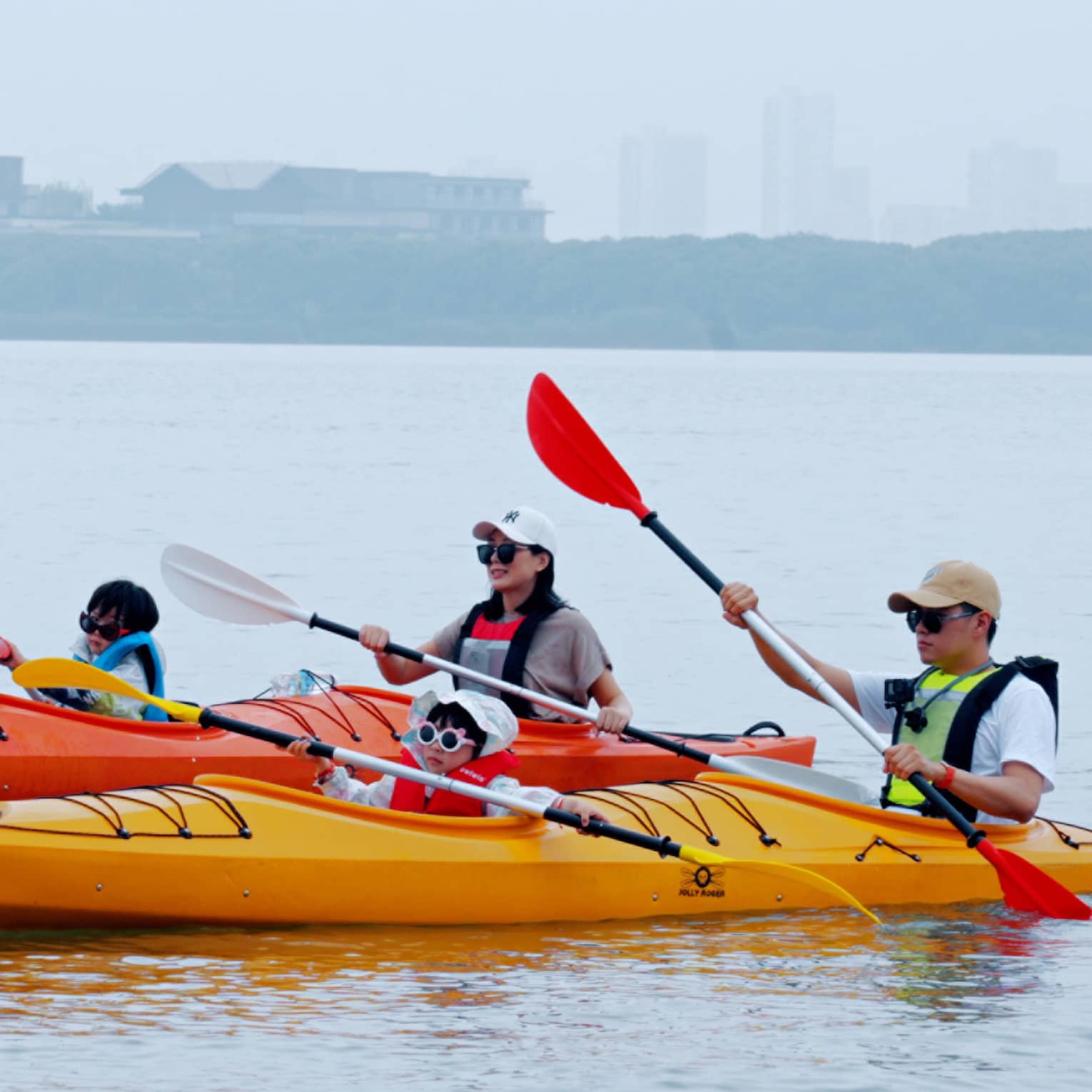 A family of four glides along serene waters, two people per kayak, distant terrain and buildings blurred by misty fog beyond.