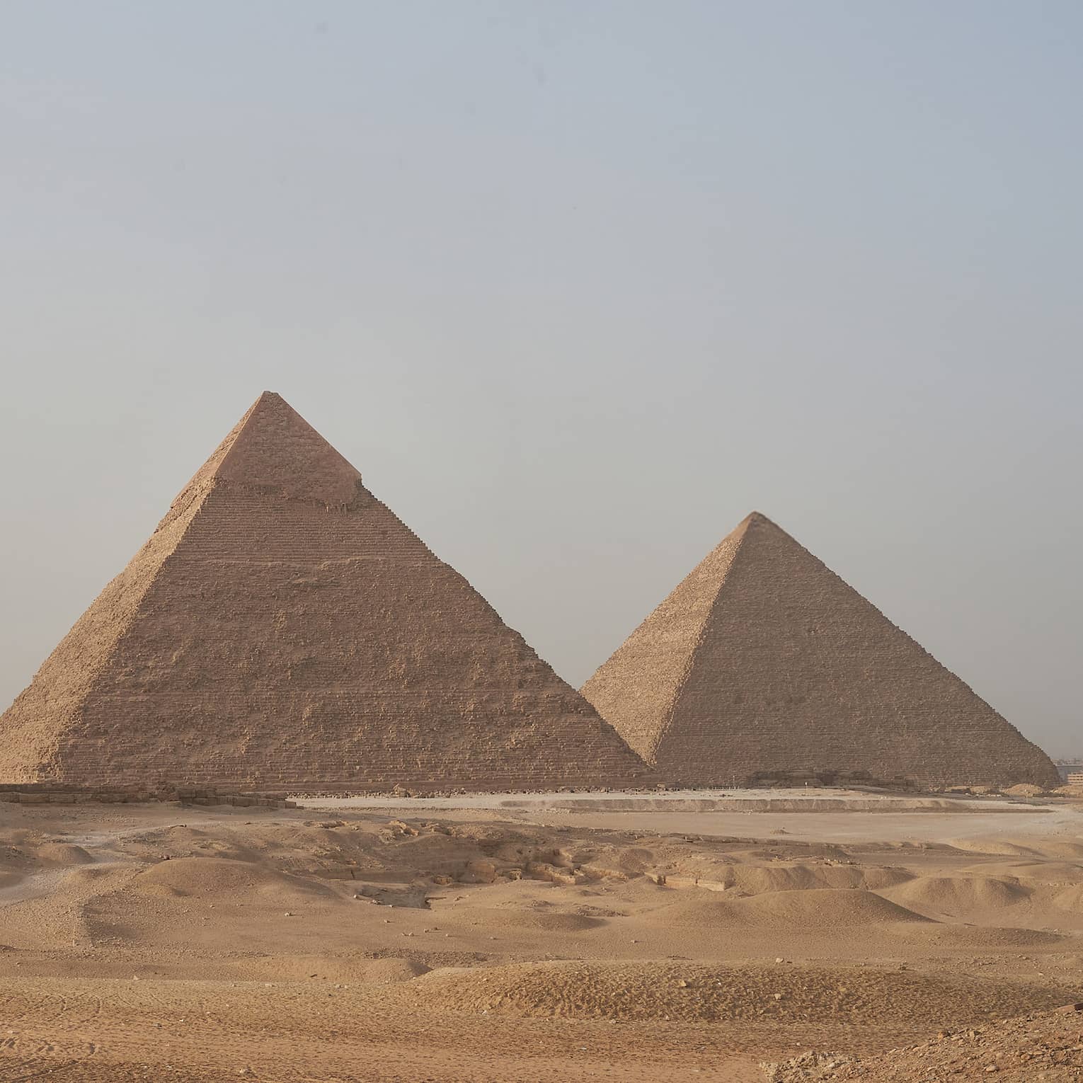 In the arid desert, a line of three towering pyramids dwarf two smaller pyramids before them, under a hazy blue sky. 